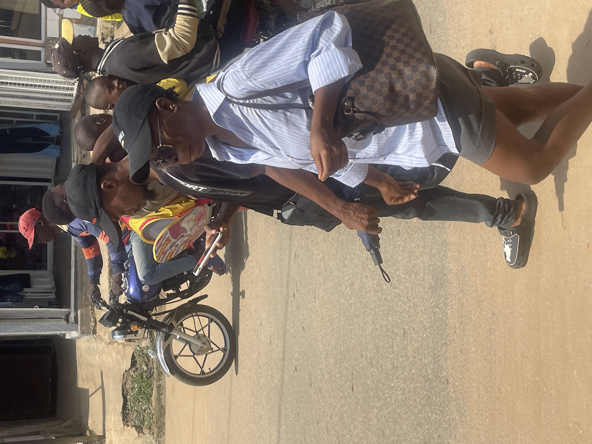 The image depicts a sunny outdoor scene in Abuja, Nigeria, focusing on a group of people on a street. In the foreground, a woman is walking to the right, wearing a light blue and white striped button-down shirt, dark grey shorts, sunglasses, and a black baseball cap. She is carrying a brown and black checkered handbag over her right shoulder and holding a closed black umbrella in her left hand. To her left and slightly behind, a man is walking, dressed in dark clothing and black sandals.
In the background, a blue motorcycle is being ridden by a man wearing a red baseball cap and a long-sleeved shirt with orange and blue stripes. He is holding a smartphone in his right hand. Several other people are visible in the background, including individuals in a yellow vest and various casual attire. They are standing and walking near buildings, suggesting a commercial or residential area. The ground is paved with asphalt and dirt. The lighting indicates it is daytime.