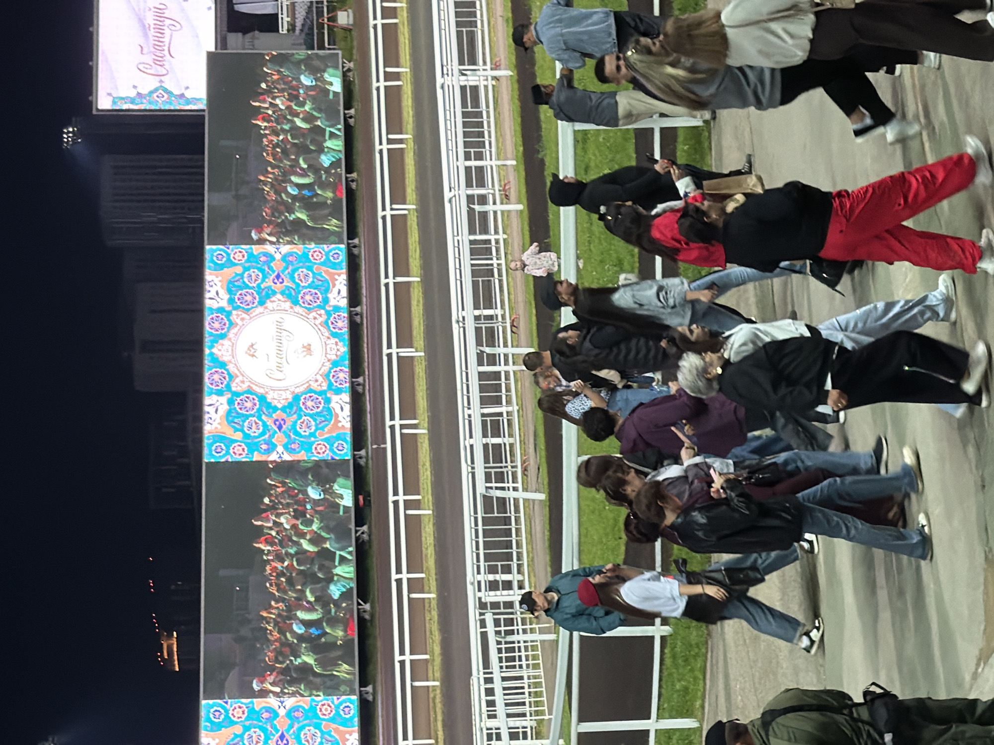 The image depicts an outdoor scene at night, likely a stadium or racetrack, as indicated by the presence of a track and spectator seating or viewing areas. A large, illuminated digital screen is prominently displayed across the top half of the image. The screen shows a crowd of people and a decorative circular emblem with script. Another, smaller screen to the right also displays a decorative emblem and script. Below the screens, a low-lying fence separates a grassy area from the track. A group of people are walking on a paved surface in the foreground, appearing to be spectators or attendees. They are dressed in casual attire. One person in the foreground is wearing a red beret and a white shirt. Another person to their right is wearing a black jacket and blue jeans. In the background, more people are visible near the fence and track, with some standing and others walking. The overall atmosphere suggests an event is taking place, possibly a performance or a gathering, given the large screens and the presence of a crowd. The lighting is artificial, coming from the screens and possibly stadium lights, casting a dark ambiance over the scene.