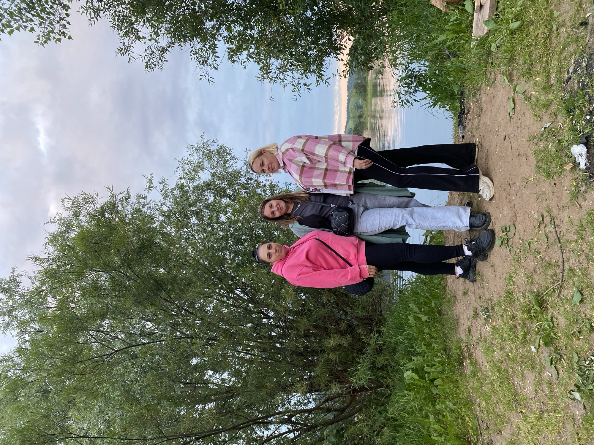 Three women are standing on a dirt and grass embankment next to a calm body of water, likely a river. The woman on the left wears a bright pink hooded sweatshirt and black leggings. The woman in the middle wears a dark gray t-shirt and light gray pants, with a green jacket draped over her shoulders. The woman on the right wears a pink and white plaid button-down shirt over black pants. They are all looking towards the camera. Behind them, a large tree with dense green foliage looms, and beyond that, the water reflects the cloudy, overcast sky. The environment appears to be natural and rural. The image was taken in the city of Noviy, Russia.