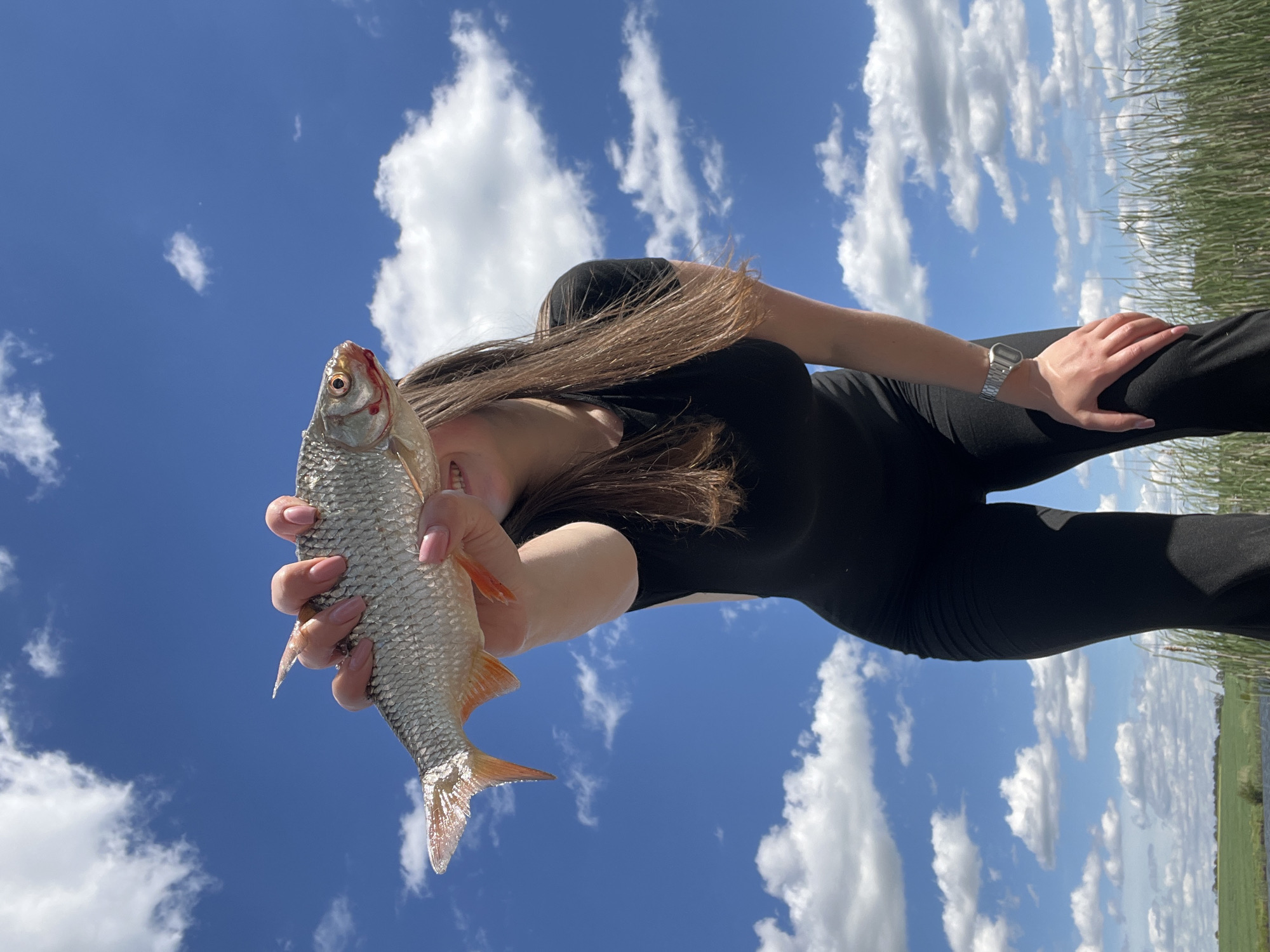 A person dressed in black is holding a fish against a bright blue sky with puffy white clouds. The person's face is partially obscured, but their light brown hair is visible. They are wearing a watch on their left wrist and have manicured nails. The fish is silver with red fins. The background also includes green grass or reeds. The image was taken outdoors during the day and it is from Rudky, Ukraine.