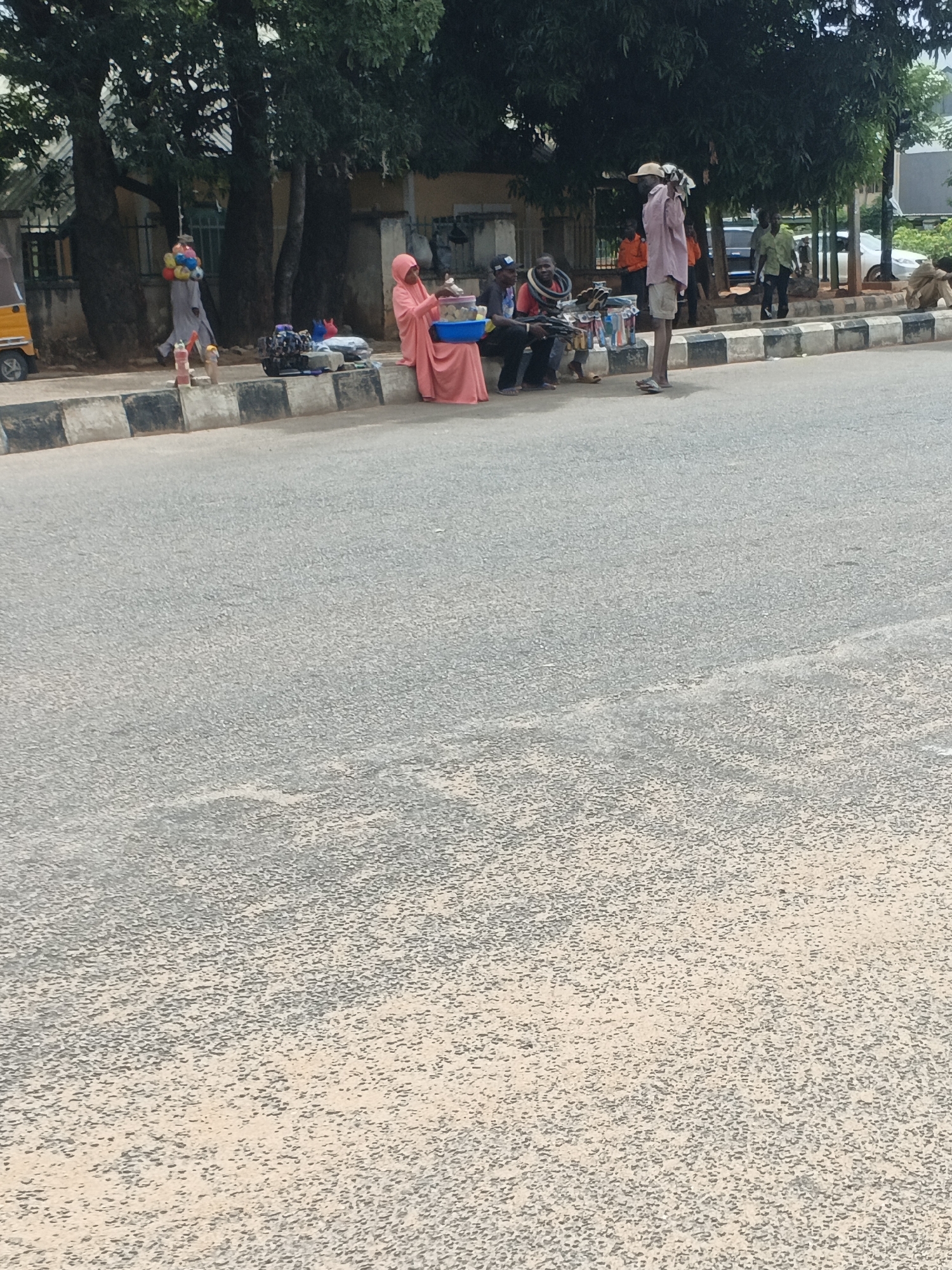 Unguwar Rimi, Nigeria: A woman in a pink hijab sits on a curb next to a vendor displaying items for sale. Two men examine the goods. A man in a hat stands beside them. Nearby, another man walks along the sidewalk carrying balloons. A yellow auto rickshaw (tuk-tuk) is parked. In the background, other pedestrians can be seen walking near buildings and trees. The foreground shows a wide asphalt street.