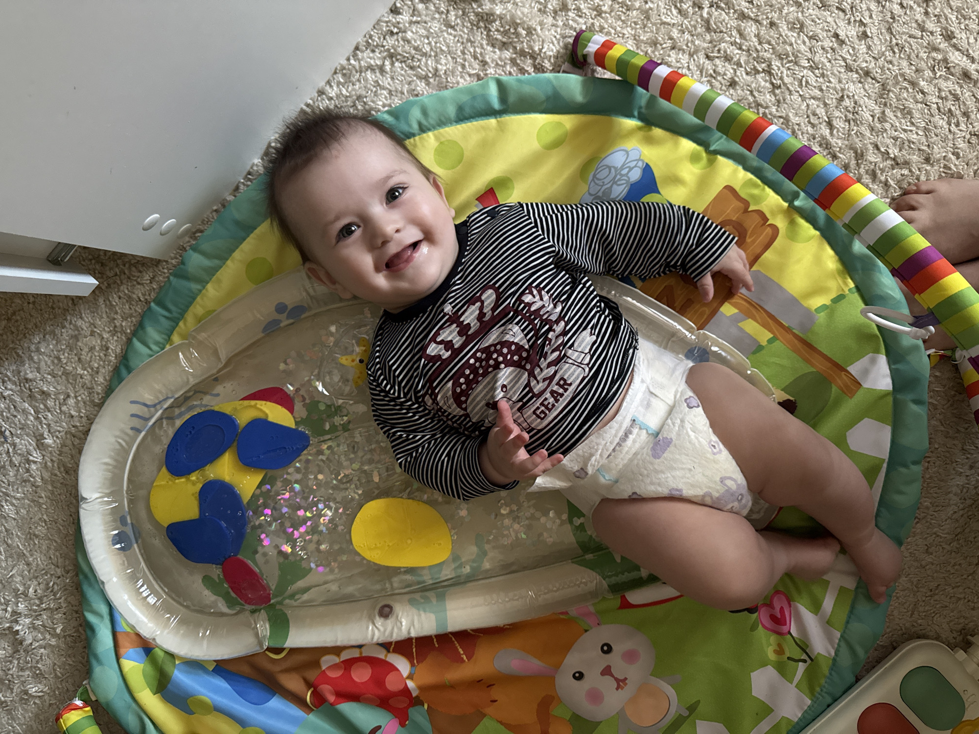A baby is lying on their back on a brightly colored play mat, smiling at the camera. The play mat is on a carpeted floor. The baby is wearing a striped long-sleeved shirt and a diaper. The play mat has a water-filled section with floating plastic shapes inside. There is a striped, soft toy arch attached to the mat. A white cabinet is visible in the background. The location is indoors, and while the image does not provide enough information to confirm the location of Kazan, Russia, the setting appears to be a child's play area in a home.