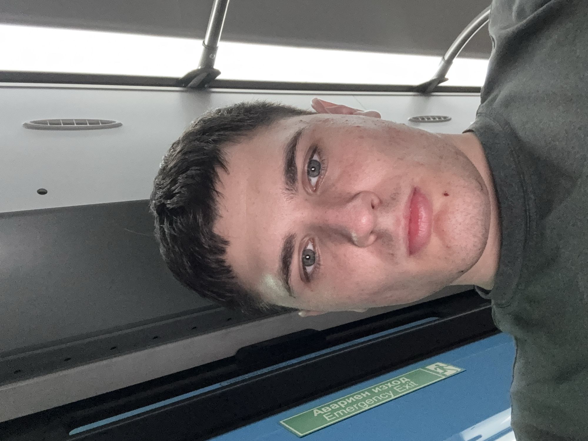 A selfie is taken from a low angle inside what appears to be public transportation, possibly a bus or train, given the visible emergency exit sign with text in Bulgarian and English, and the overhead lighting and vents. The subject is a young Caucasian male with short, dark hair, light-colored eyes, and fair skin with some blemishes. He is wearing a dark grey crew-neck t-shirt. He is looking directly at the camera with a neutral expression. The background is blurred, showing a blue panel with the green emergency exit sign and a grey interior with a long, bright light fixture. There is no indication of the city of Stara Zagora, Bulgaria, in the image itself.