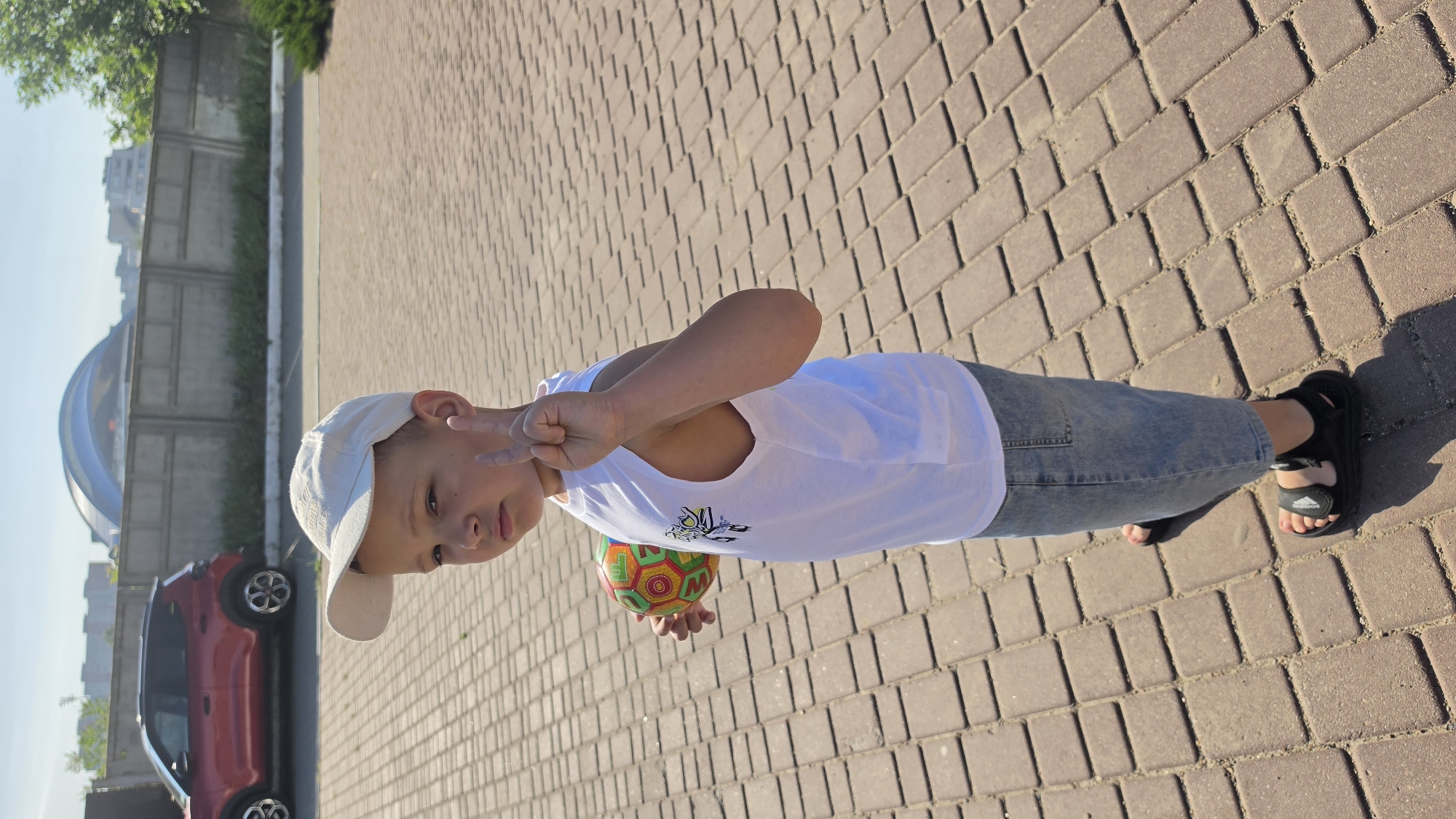 A young boy wearing a white baseball cap, a white sleeveless shirt, and denim shorts is standing on a paved walkway. He is holding a colorful ball in his left hand and making a peace sign with his right hand. In the background, a red SUV is parked near a concrete fence. Beyond the fence, there are buildings and a large, arched structure. The scene appears to be outdoors in a city.