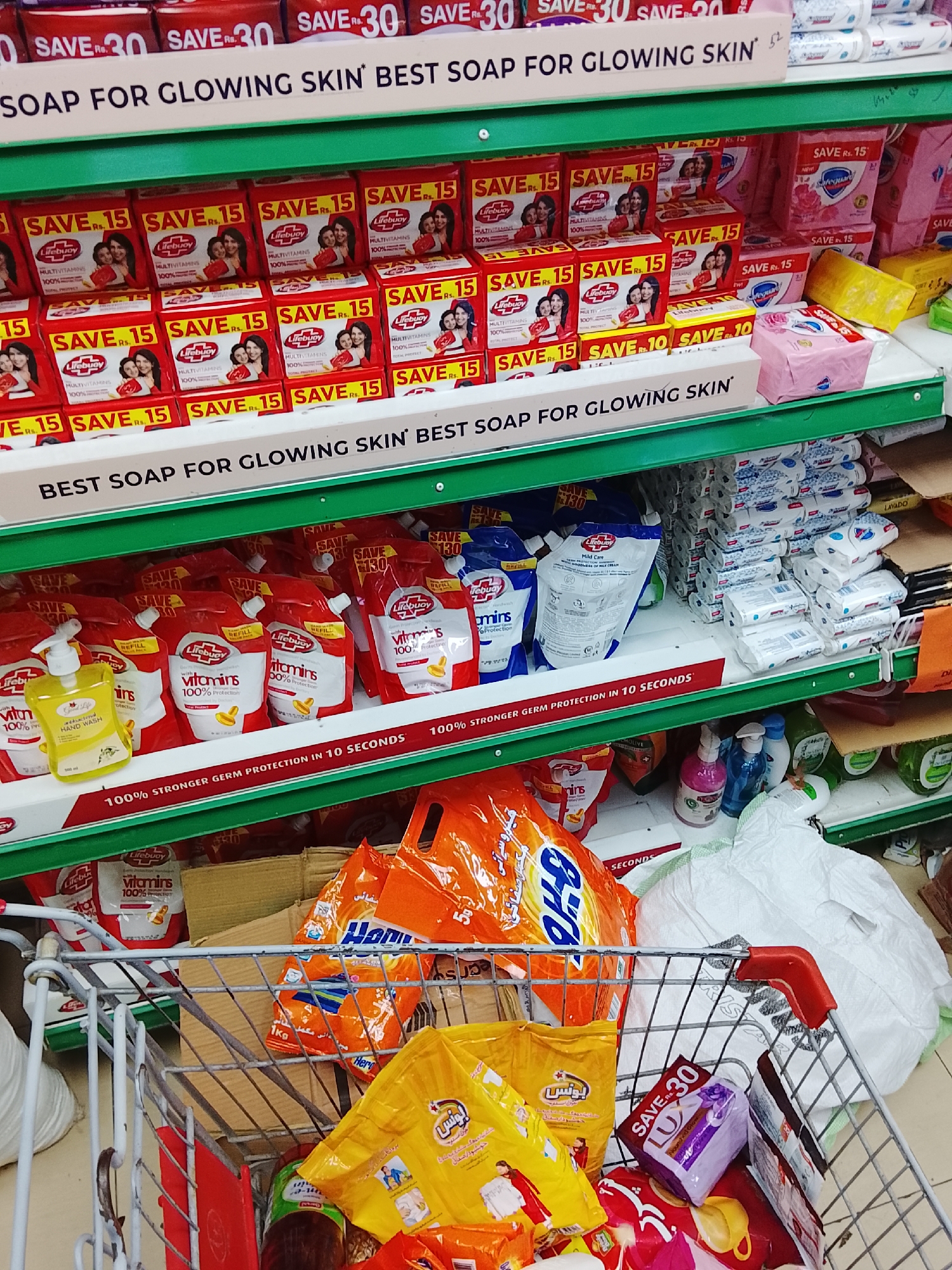 The image depicts a shopping scene inside a store located in Gulgasht Colony, Pakistan. The focus is on a metal shopping cart containing various household and personal care items, including detergents and soaps from brands such as Lifebuoy, Bonus, and Lux. Behind the shopping cart are shelves stacked with similar products. The shelves are stocked with Lifebuoy hand wash, Lifebuoy refill pouches, and various types of soap. The price tags are visible, indicating savings on certain products. The store's aisle is visible.