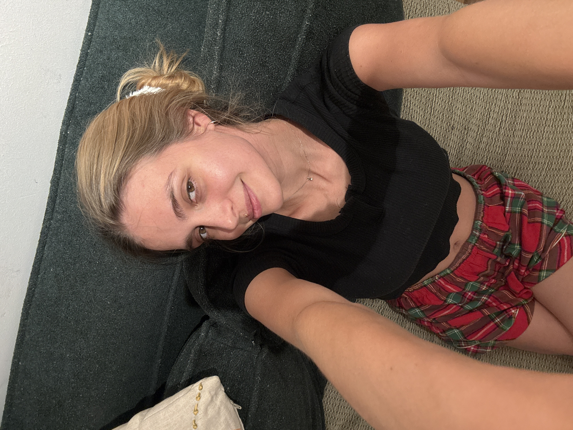 A young woman with blonde hair in a messy bun is lying on a dark gray couch, wearing a black ribbed crop top and red plaid shorts. She is taking a selfie, holding the camera with one arm extended towards the viewer. Her face is slightly tilted towards the camera. She has a piercing on the left side of her nose and a silver necklace. The background includes the couch, part of a white wall, and a textured beige floor.