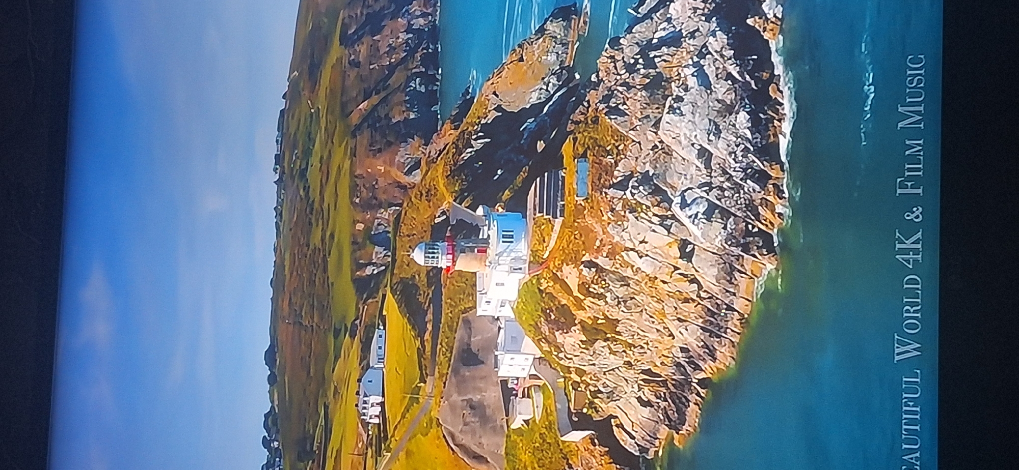 The image shows an aerial view of a lighthouse on a rocky cliff overlooking the sea. The lighthouse is white with a red band and a white lantern room. There are several other white buildings surrounding the lighthouse, suggesting it is part of a larger complex. The cliffs are rugged and covered in green vegetation. The sea is a vibrant blue, with some whitecaps visible. In the background, there are rolling hills with more vegetation and a few scattered houses. At the bottom of the image, there is white text that reads "BEAUTIFUL WORLD 4K & Film Music." The scene is bathed in bright sunlight, indicating a clear day. There is no presence of the city of Bila Tserkva, Ukraine in this image.