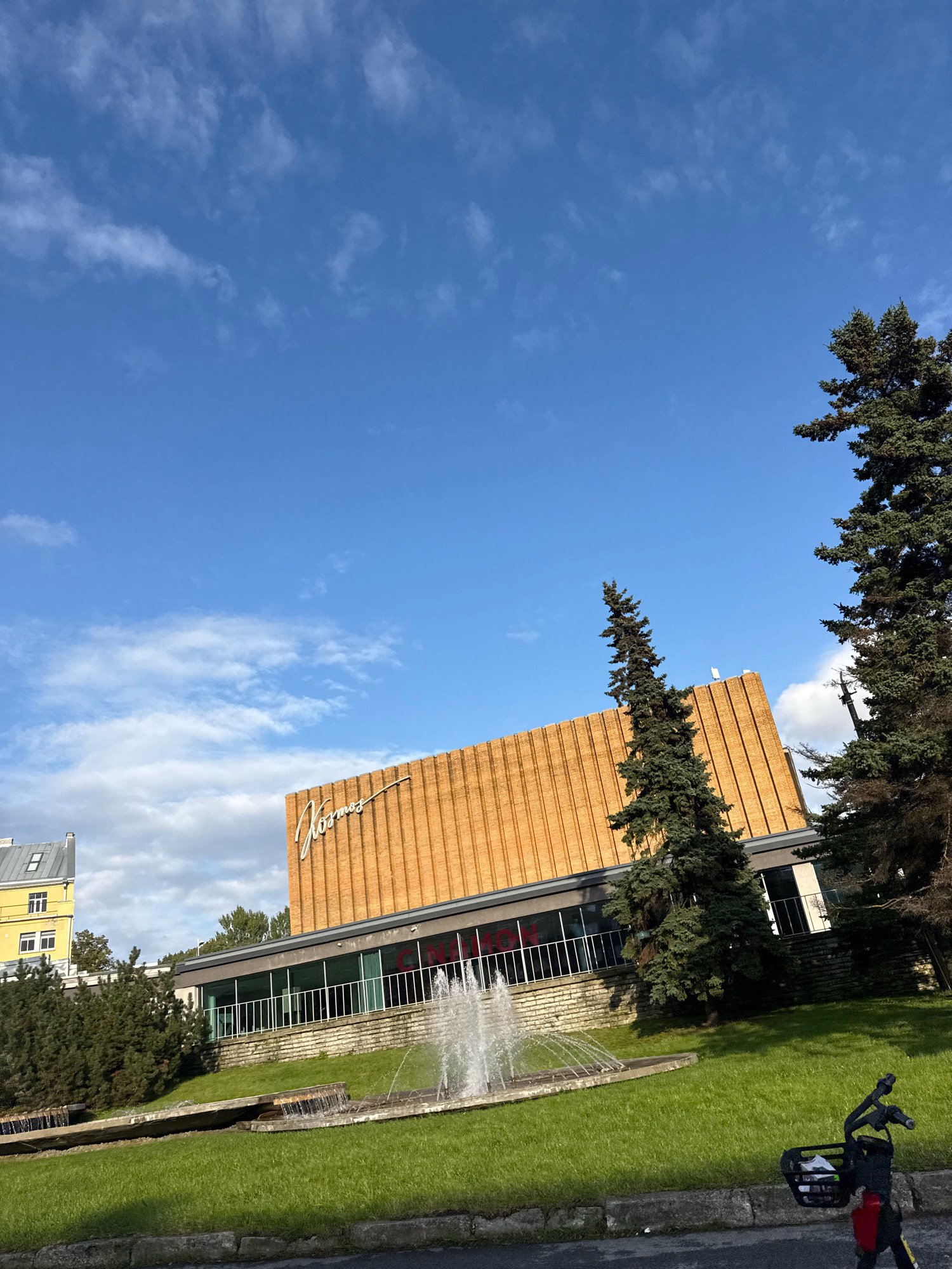 This image depicts a daytime exterior view of the Kosmos cinema in Tallinn, Estonia. The cinema is characterized by its distinct orange, vertical ribbed facade with a white, cursive "Kosmos" sign. In front of the cinema, a terraced fountain with cascading water is visible, surrounded by a well-maintained green lawn. Tall evergreen trees flank the building and the fountain area. To the left of the cinema, a yellow apartment building with multiple windows is partially visible, suggesting a mixed urban environment. The sky above is a clear blue with scattered white clouds. In the foreground, the handlebars and basket of a red and black electric scooter are visible, positioned on a paved surface.