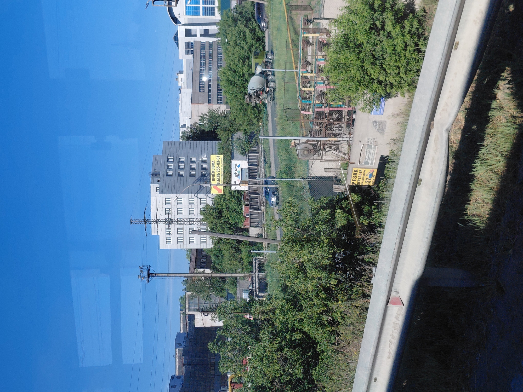 This is an eye-level, medium shot of an industrial area under a clear blue sky. In the foreground, a silver guardrail runs horizontally across the frame, casting a shadow on the dry grass and weeds below. Beyond the guardrail, an assortment of industrial buildings, some with brick exteriors and others with white siding, are visible. Electrical towers and power lines crisscross the scene. A cement mixer truck is driving on a road, with a few cars parked or driving in the distance. There are also several trees and lush green vegetation scattered throughout the area, contrasting with the man-made structures. A yellow sign with Russian text is visible on the left side of the image. The overall impression is one of an active, albeit somewhat dated, industrial zone.