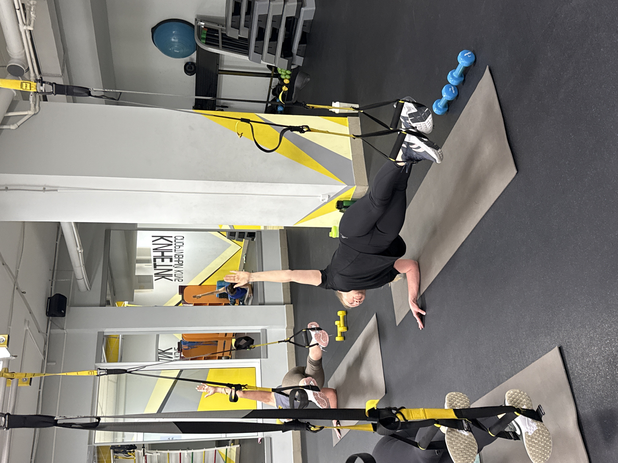 The image shows two women using TRX suspension straps in a gym. One woman is performing a handstand with her feet secured in the TRX straps, while the other is on her back with her feet secured in the TRX straps. The gym setting includes mats, weights, and a mirrored wall with the name "KINE LNK" and what appears to be its Cyrillic transliteration. Based on the name, the location is very likely in the city of Chaikovsky, Russia.