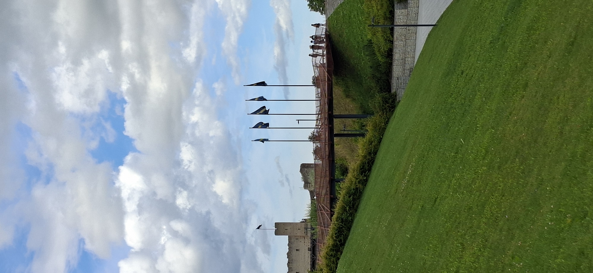 The image depicts the Rakvere Castle in Rakvere, Estonia, under a partly cloudy sky. In the foreground, a lush green lawn slopes upwards, leading to a modern, reddish-brown elevated walkway. This walkway, supported by dark pillars, is lined with several flagpoles flying yellow and blue flags, with one black flag visible on a tower to the left. A group of people are walking on the elevated walkway. Behind the walkway, the stone walls and towers of Rakvere Castle are visible, suggesting historical significance. The overall scene is a blend of natural landscape and historical architecture with modern amenities.
