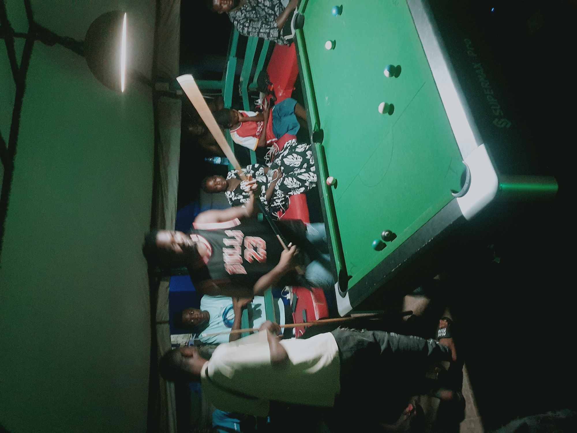 A dimly lit indoor scene in Lokoja, Nigeria, shows a group of people gathered around a green felt pool table with multiple billiard balls scattered on its surface. Some individuals are seated on benches near the table, while others are standing. One man, wearing a Chicago Bulls jersey, appears to be aiming with a pool cue. A light fixture illuminates the pool table from above. A logo "SUPERMAX PRO" is visible on the side of the table. The surrounding environment seems to be a bar or recreational area.