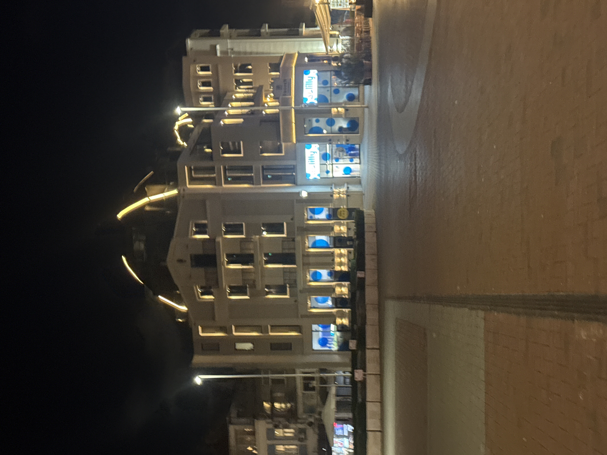 The image depicts a building at night in Varna, Bulgaria. The building has multiple stories with windows, some illuminated. There is a sign for "Lilly" on the building facade. Streetlights and decorative lights are visible. The ground appears to be paved, and a sidewalk is present. The night sky is dark.