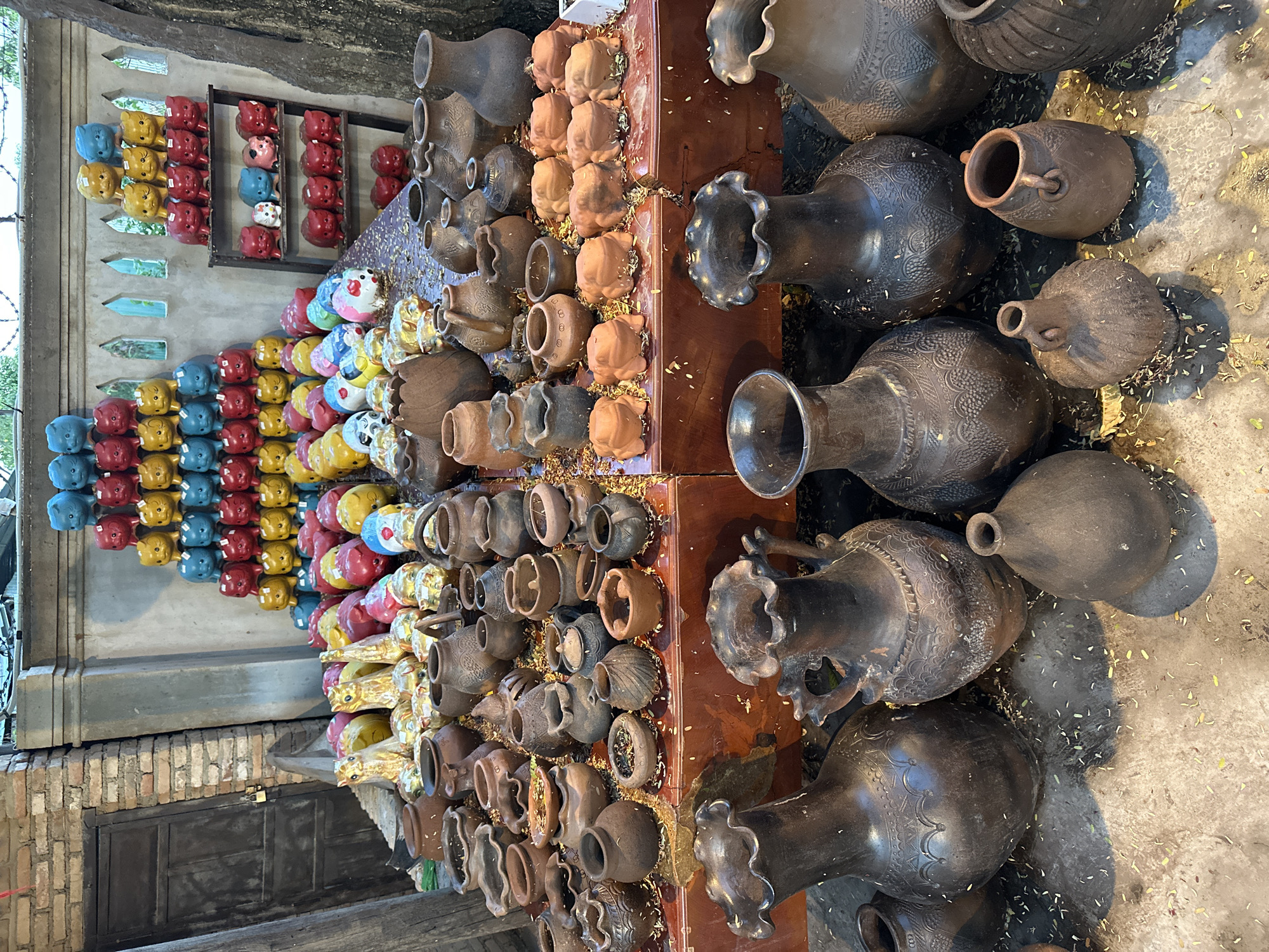 The image depicts an outdoor market stall in Nha Trang, Vietnam, specializing in pottery and ceramic figures. In the foreground, a collection of dark, earthen-toned pottery vases and jars of various sizes are displayed. Some have ornate patterns etched into their surfaces, while others are simpler in design. Scattered on the ground around the pottery are dried leaves and petals.
Behind the pottery, on a tiered display area, are rows of brightly colored ceramic figures, predominantly in the shape of pigs and cats, stacked in a pyramid-like formation. These figures are primarily in shades of blue, red, and yellow, with some featuring details like eyes and ears. Further back, against a wall with decorative cutouts, more ceramic figures are visible, along with some golden animal figurines. The background also shows elements of brickwork and tree trunks, suggesting a natural, rustic setting. There are no people visible in the image.
