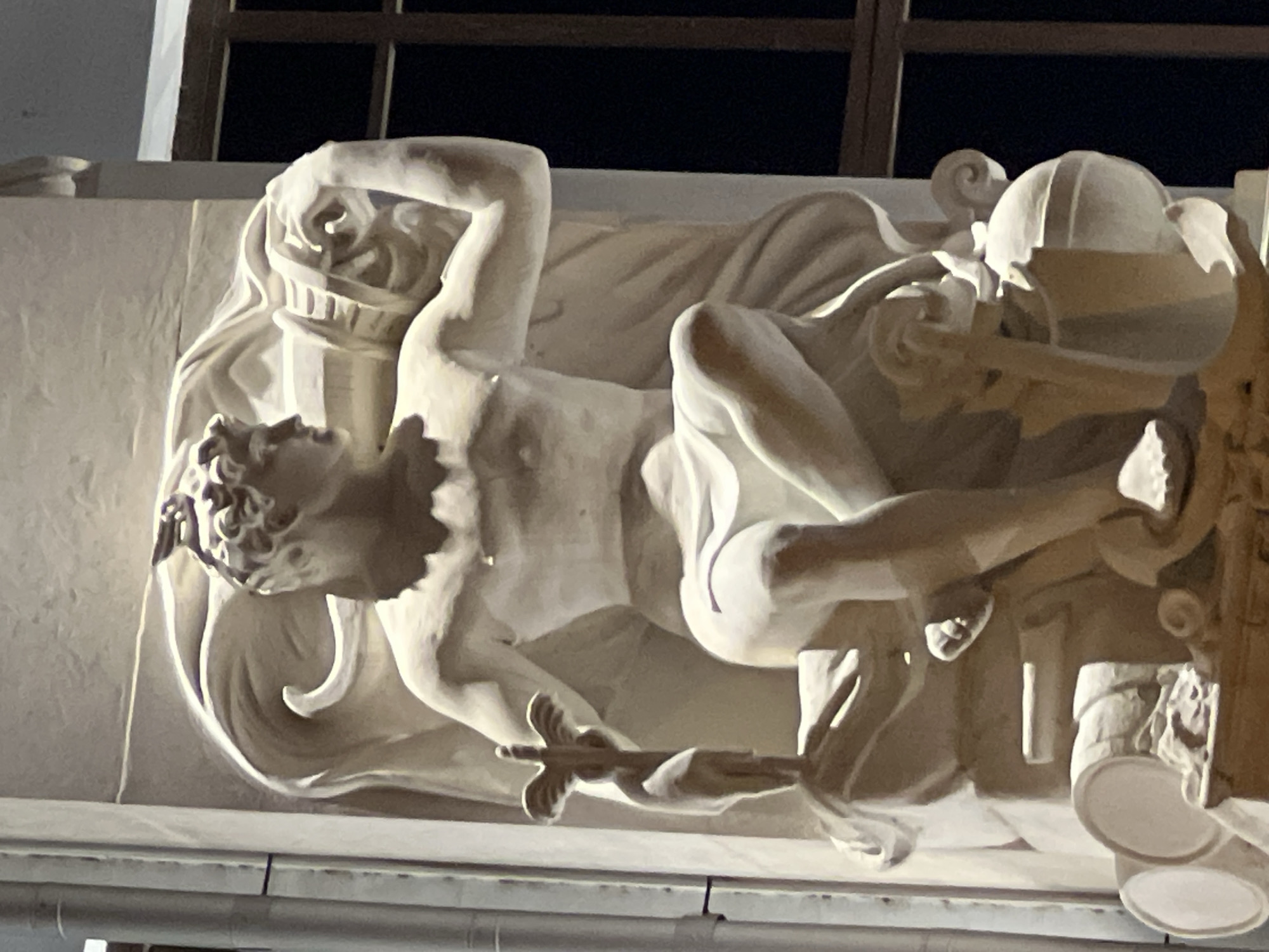 The image shows a detail of a white stone sculpture of a male figure. The figure is seated, with his left leg bent and his right leg extended. He is holding a staff in his left hand and a cylindrical object in his right hand, which is raised to his head. The sculpture is ornate, with swirling drapery behind the figure and various objects at its base, including what appear to be barrels and a globe. The sculpture is illuminated by artificial light, casting shadows and highlighting the texture of the stone. In the background, a dark window frame with mullions is visible. The sculpture is located on a building in Krasnodar, Russia.