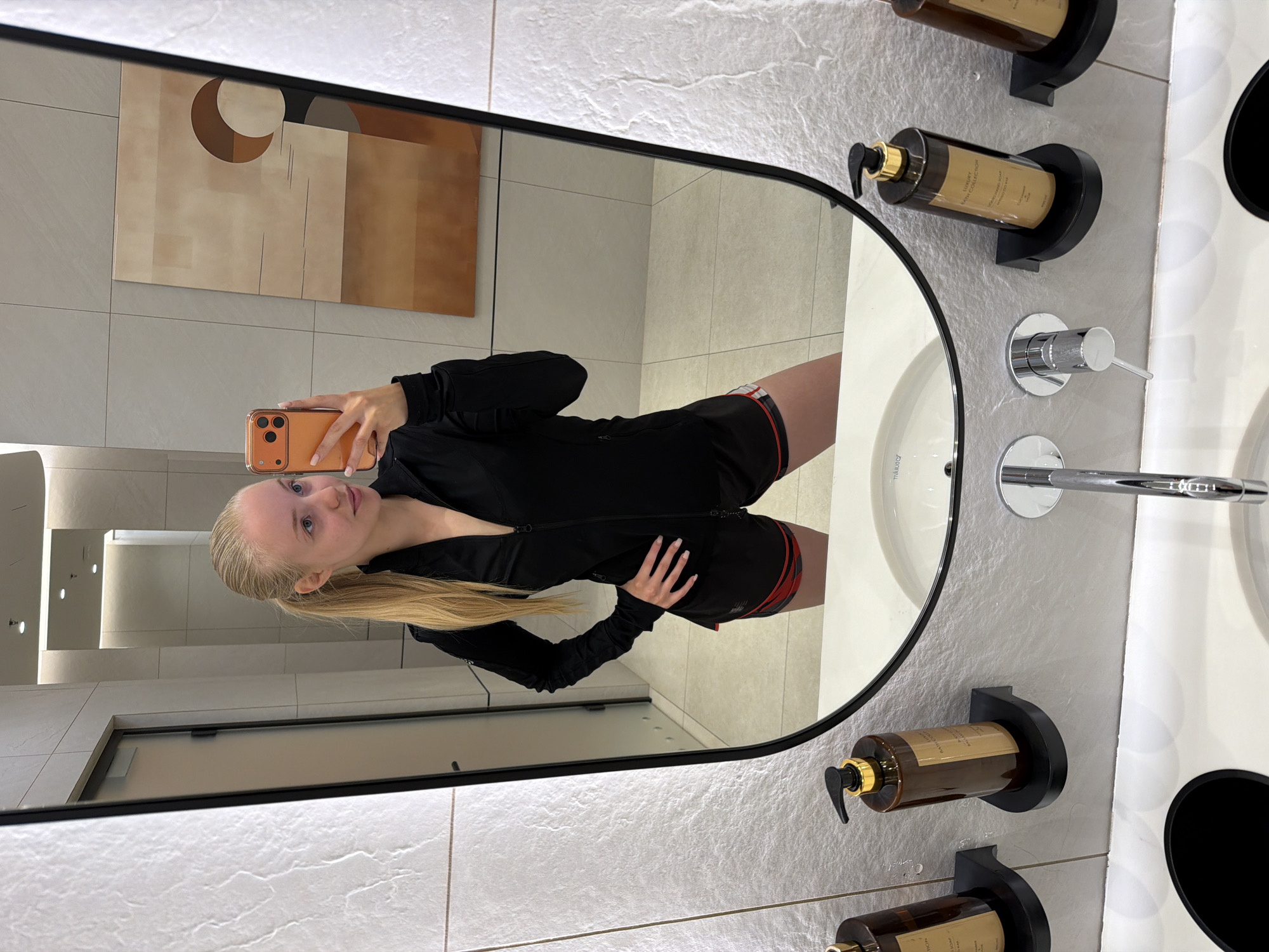 The image is a selfie of a blonde woman wearing a black athletic jacket and red/black shorts, taken in a mirror in a restroom. The reflection in the mirror shows a neutral colored abstract painting on the wall behind her. The restroom has textured, light-colored walls and a white countertop with a sink. On the counter are several brown soap dispensers and chrome faucet fixtures. The location is Unknown, Unknown.