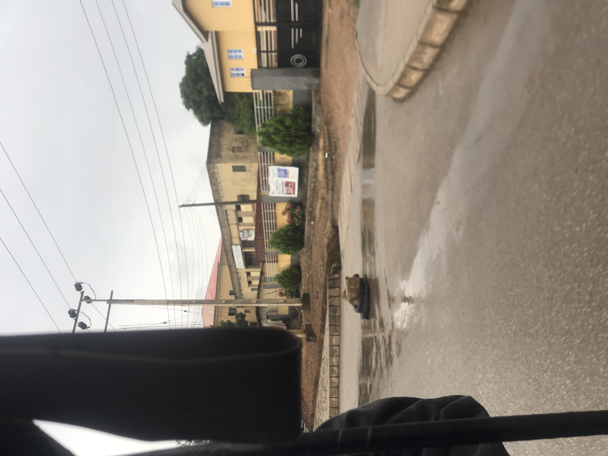 The image displays an overcast street scene, likely in New Stadium, Nigeria, with clear indications of recent rainfall. The perspective appears to be from inside a vehicle, with a dark portion of the vehicle's interior visible at the bottom of the frame.
The central subject is a wet, paved road with numerous puddles reflecting the dull sky. To the right, a raised curb and sidewalk run alongside the road. On the left, an unpaved shoulder features sparse green bushes and what appears to be a small, light-colored object resting in a puddle on the road.
In the background, several multi-story buildings with light yellow or beige facades are visible, some with dark security gates. Lush green trees are scattered among the buildings and in the distance. Overhead, multiple power lines crisscross the sky, supported by utility poles. A white banner or sign, bearing illegible text or graphics, is attached to one of the buildings in the mid-distance.
The time of day is not clearly discernable but is likely daytime due to the ambient light, though obscured by heavy cloud cover. The wet surfaces and puddles are the most notable weather detail, suggesting it has either just rained or is still drizzling. There are no people visible, and no specific activity is taking place, creating a quiet and somewhat desolate atmosphere.