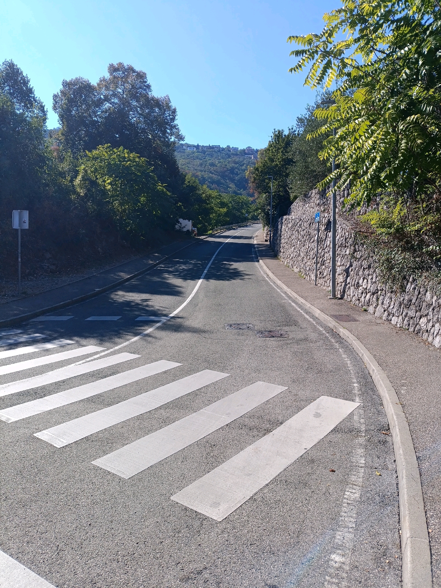 The image shows a clear day in Opatija, Croatia. A paved road curves to the right, with a clear blue sky overhead. In the foreground, a pedestrian crossing is marked with white stripes on the asphalt. To the right of the road, a stone wall borders a sidewalk. Lush green trees and vegetation line the left side of the road and the slope above the stone wall. In the distance, houses are visible on a hillside. A lamppost stands on the sidewalk next to the stone wall. A small white sign with a red symbol is mounted on a pole on the left side of the road, near the pedestrian crossing.