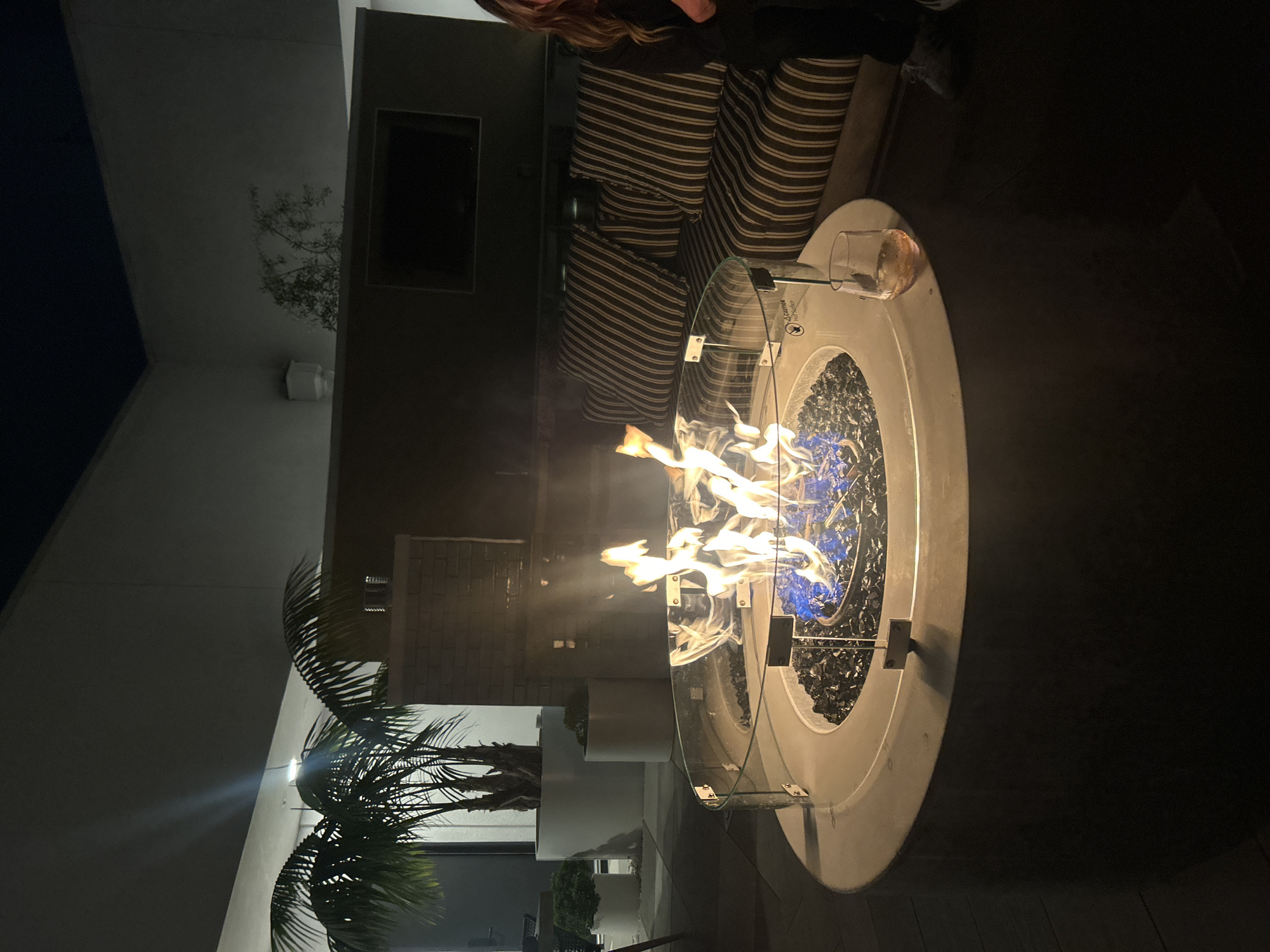 The image displays an upscale outdoor patio or rooftop terrace at night, located in a modern building in Los Angeles, California, United States of America.
In the foreground, a circular, light-colored concrete fire pit is active, emitting blue and orange flames from a bed of black fire glass. A clear glass wind guard, comprised of four panels held by metal clips, surrounds the burning area, and a "CAUTION Hot Surface" label is visible on the fire pit's rim. A clear, stemless glass holding a light amber-colored liquid rests on the edge of the fire pit closest to the viewer.
To the right, a segment of an outdoor sofa with dark brown and light beige vertical stripes on its cushions is visible. A person with long brown hair, wearing dark clothing, is partially observed to the right of the sofa, with the right side of their face, shoulder, and a light-colored shoe visible near the base of the fire pit.
The background features modern, light-colored walls and darker architectural elements, including a rectangular recess. Two large palm trees are situated in tall white planters on the left side of the scene, with smaller potted plants positioned behind them. The ground is surfaced with dark-colored tiles or planks. Overhead lighting fixtures provide supplementary illumination to the setting. A partial brick column is visible to the left of the sofa.