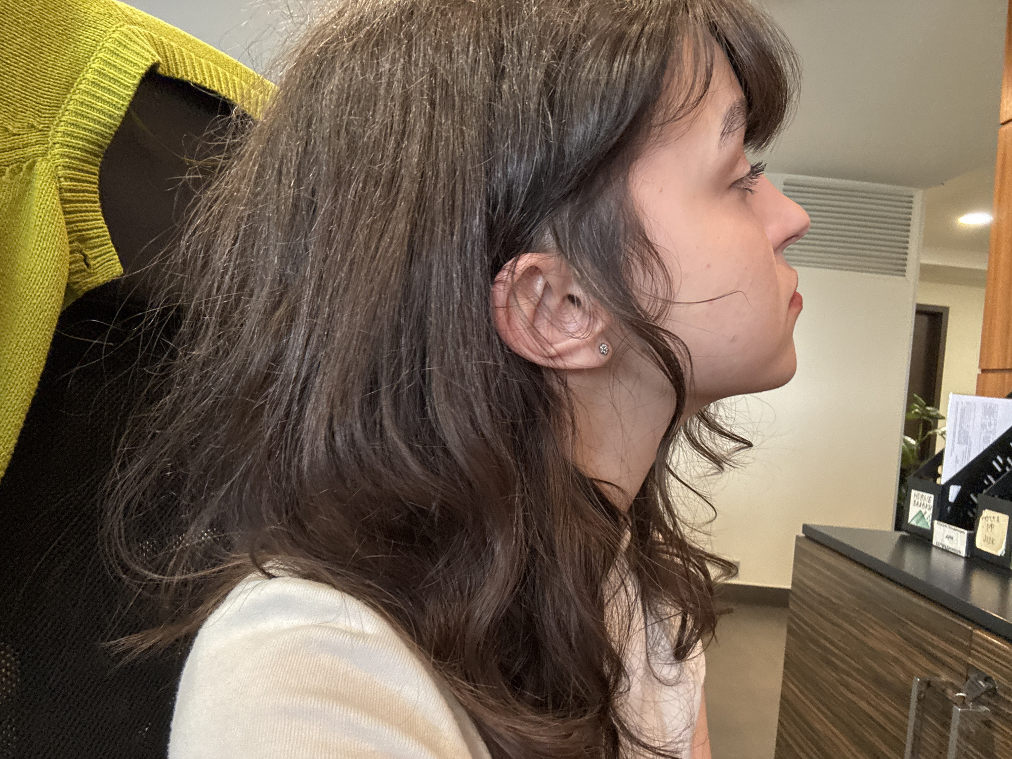 A side-view medium shot shows a woman with brunette wavy hair. She is wearing a light beige top and a lime green sweater is partially visible in the upper left of the frame. She has fair skin, dark eyelashes, and a small stud earring in her left ear. She is looking towards the right of the frame. A dark chair is behind her. In the background, a desk with office organizers and a cabinet are visible. The scene is illuminated by overhead lighting. The environment suggests an indoor office space, though there is no evidence to confirm the scene's location in Saint Petersburg, Russia.