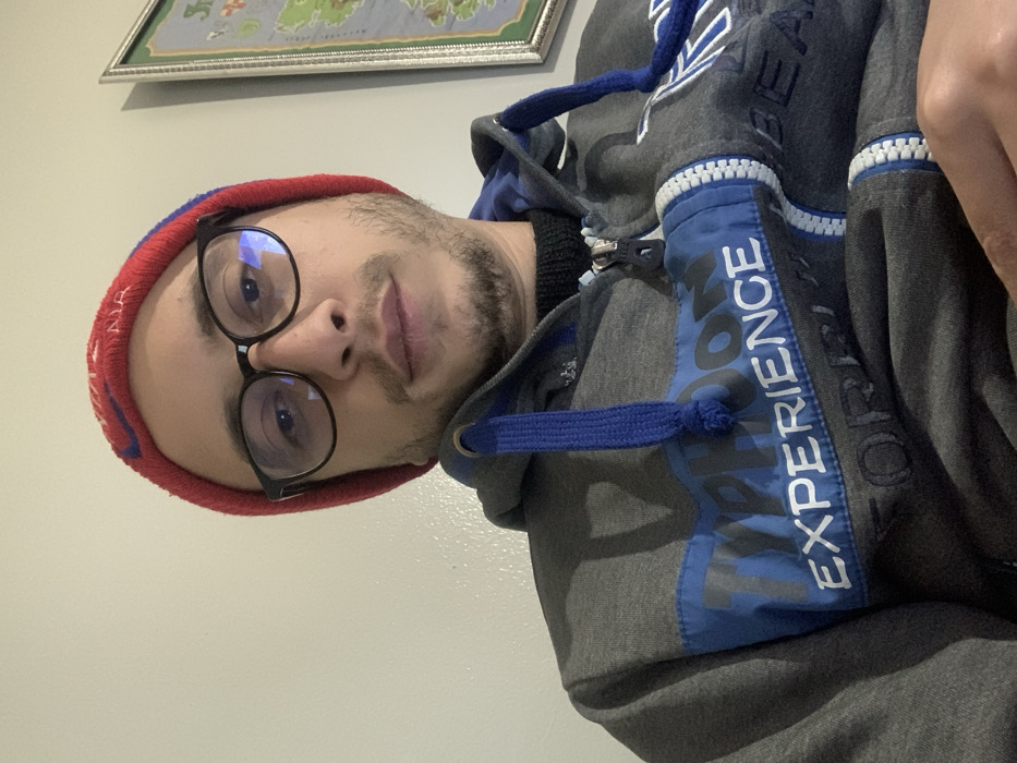 A man wearing a red beanie and black-framed glasses is looking at the camera. He has a light beard and mustache. He is wearing a grey hooded sweatshirt with blue accents and the word "EXPERIENCE" printed vertically on the front. A framed map hangs on the wall behind him. The image is rotated counter-clockwise.