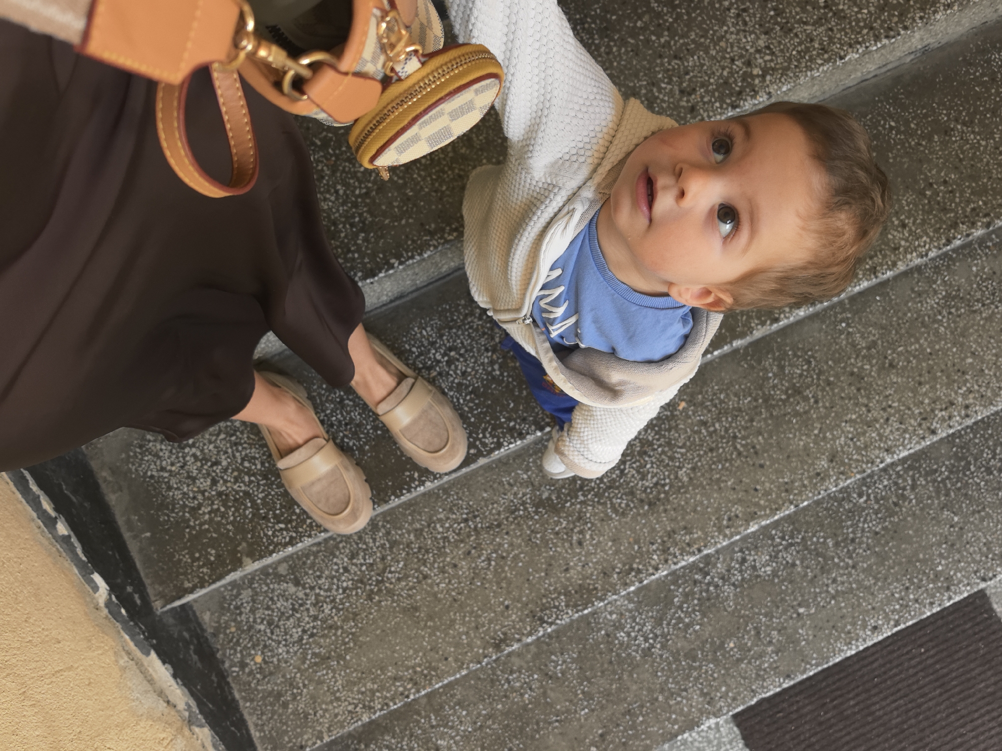 A high-angle shot shows a woman and a young child standing on concrete stairs. The woman, partially visible, wears a long brown skirt and beige suede loafers. She holds a brown leather handbag and a smaller coin purse accessory. The child, wearing a white hooded sweater and a blue shirt, looks upwards with a curious expression. The environment includes concrete stairs with some discoloration and a glimpse of a dark rug at the bottom of the stairs. The location is Stara Zagora, Bulgaria.
