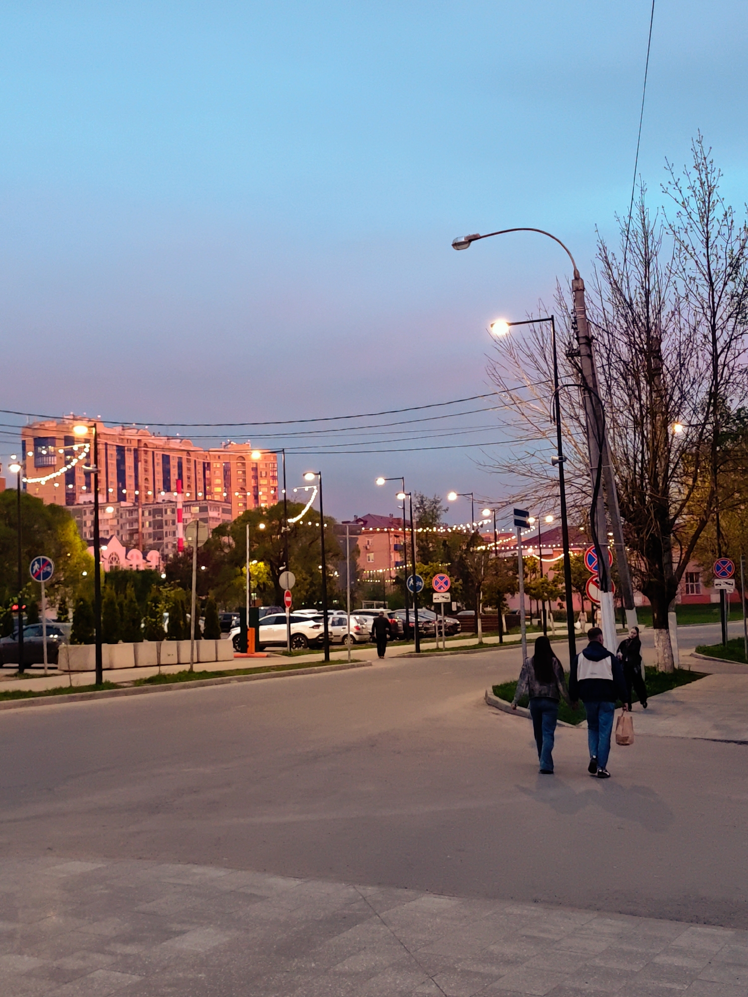 The image shows a street scene in Dmitrov, Russia, at dusk. The sky is a gradient of blue and pink. An orange-lit building with string lights is visible in the background. Street lights illuminate the road and surroundings. Several cars are parked along the street. Two people are walking away from the viewer on the sidewalk. A utility pole with wires and bare trees are on the right side of the image. Various street signs are visible along the road and sidewalks.