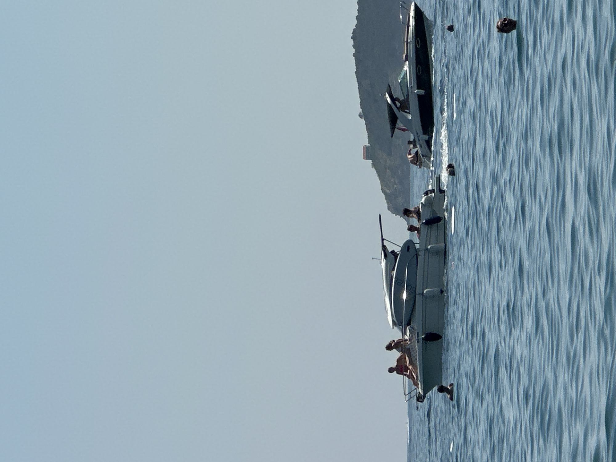 Two motorboats are on the water under a clear sky. The boat on the left has several people on board. The boat on the right is moving, with people visible inside. In the background, a hilly island with a small building on top is visible. Several people are in the water, swimming. The scene appears to be a typical summer day at a coastal location. The location is likely near Boreti, Montenegro, given the geographical features.