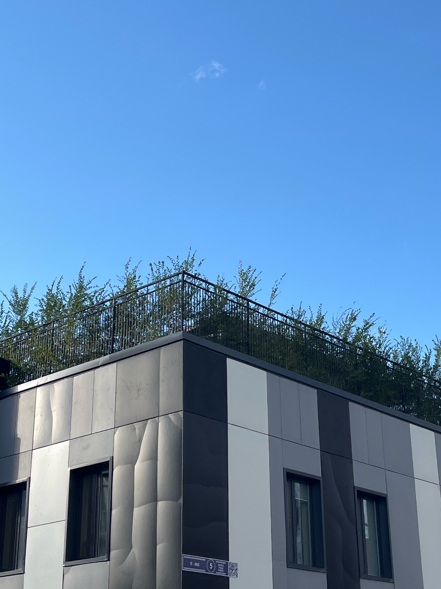 The image shows the exterior of a modern building with a black fence and lush green trees on its rooftop. The building's facade is a mix of gray and white panels, some with a textured, wavy pattern. There are several rectangular windows visible. The sky above is a clear, bright blue with a small, wispy cloud. A street sign with the number "5" is partially visible on the lower right side of the building. The architectural style suggests it is located in a contemporary urban environment.