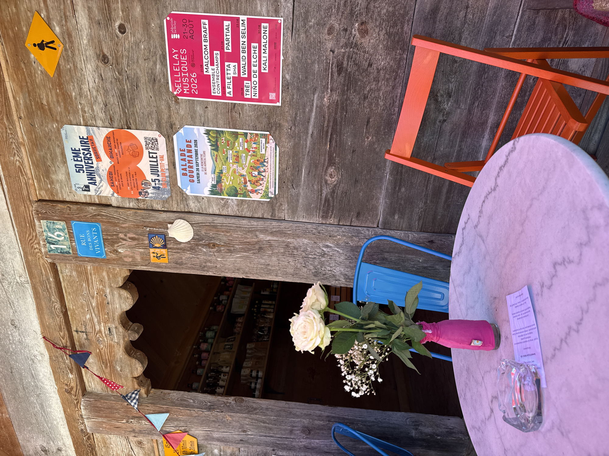 The image depicts an outdoor seating area in front of a wooden building. In the foreground, a round marble-top table is set with a glass ashtray and a pink vase containing white roses and baby's breath. An orange folding chair is positioned to the right of the table, and a blue chair is visible to the left.
The wooden building facade features several posters. One poster announces the "50 EME ANNIVERSAIRE SORBETAN" on July 4-5. Another poster, titled "BALADE GOURMANDE," advertises an event on September 26, 2026, featuring a meeting with agricultural producers. A third, pink poster lists "BELLELAY MUSIQUES 2026" with a schedule of performances from August 21-30.
Above the entrance, a sign reads "16" and below it, "RUE DES BONS VIVANTS." Further decoration includes a string of small fabric flags and a yellow diamond-shaped sign with a hiker icon. Two small square tiles, one depicting a pilgrim and the other a shell, are also present. A white shell ornament is affixed to the wooden wall.
Inside the building, shelves lined with various bottles and jars are visible through an open doorway. The overall scene suggests a relaxed outdoor setting, possibly a cafe or a shop with seating, located in Petit-Val, Switzerland, as indicated by the posters.