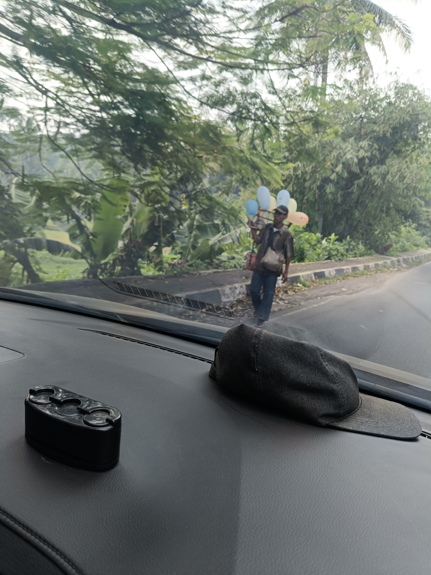 A street vendor carrying balloons walks along a paved road in a rural area, possibly near Pandeglang, Indonesia. The vendor, a person of unspecified gender, is wearing a dark shirt, blue pants, and a dark cap. They are carrying a bundle of multi-colored balloons. A bag is slung over their shoulder. The road is bordered by a sidewalk and lush green vegetation, including trees and what appear to be banana plants. In the foreground, on the dashboard of a vehicle, lies a black baseball cap and a black object with multiple circular indentations. The scene is viewed from inside the vehicle.
