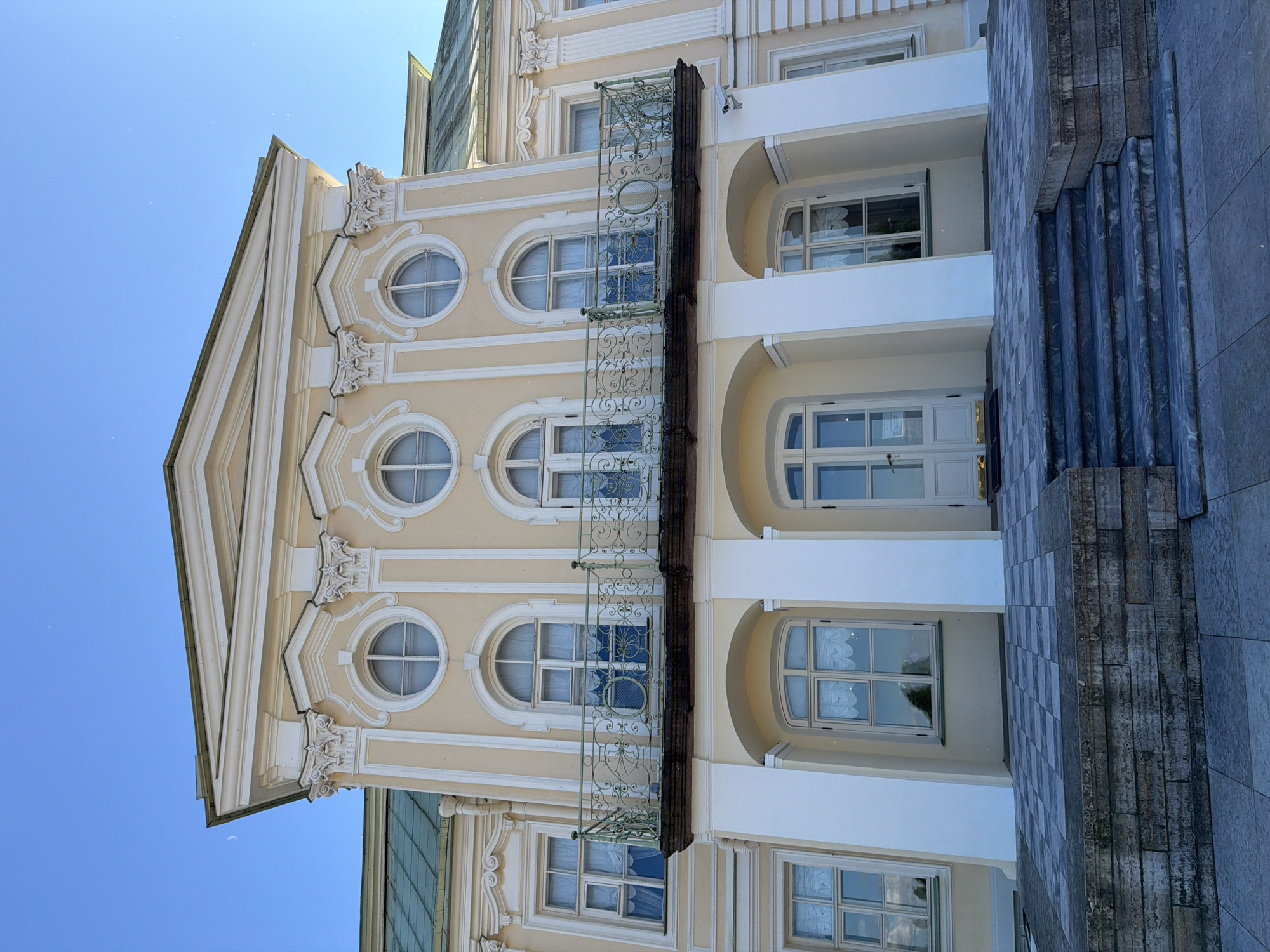 The image shows the facade of a pale yellow building with white trim, likely a palace or stately home, under a clear blue sky. The architecture features classical elements such as columns, ornate pediments, and rounded and arched windows. A balcony with an intricate green railing is supported by large white pillars that form an arched entranceway. The entrance has double doors, and windows on either side are visible. A paved walkway with a checkered pattern leads up to a set of stone steps in front of the entrance. The building appears well-maintained, and the setting suggests an outdoor, daytime scene. There are no people visible in the image.