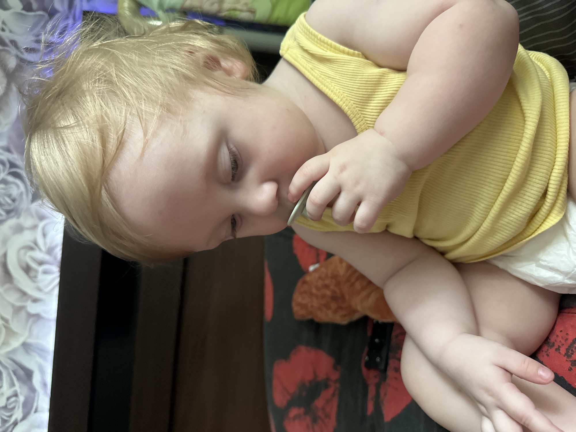 The image shows a close-up of a baby with blonde hair and fair skin, lying on their side. The baby is wearing a yellow ribbed tank top and a diaper. Their eyes are closed, and they appear to be sucking on a pacifier or a small object. The baby's left arm is resting on their body, and their right arm is bent, with their hand near their mouth. There is a stuffed toy peeking out from under the baby's arm. The background is blurred, but it appears to be an indoor setting with patterned fabric. One section of the background shows a white floral pattern on a dark surface, and another shows a dark brown surface. The lighting suggests it is daytime. The overall mood is peaceful and sleepy.