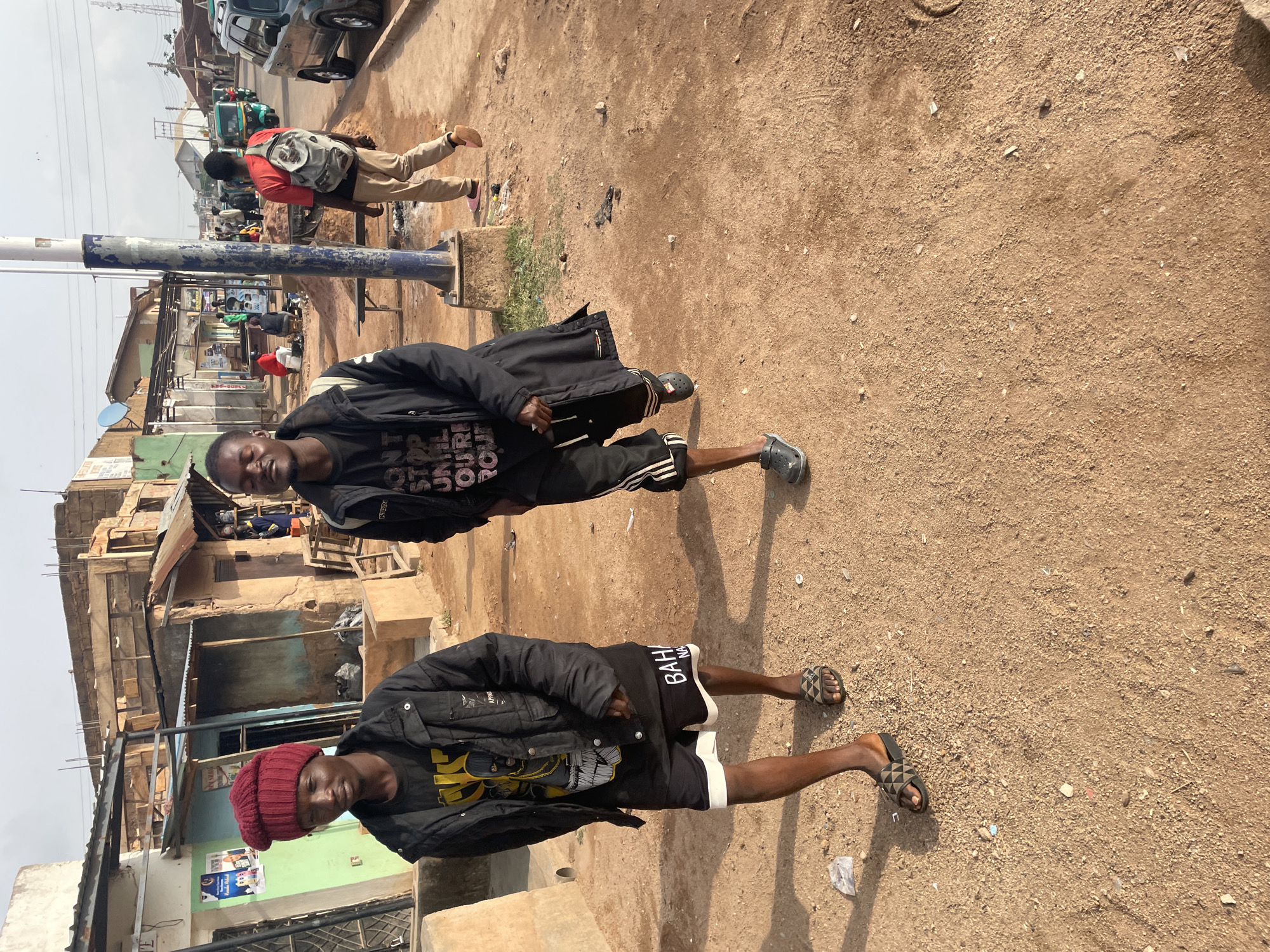 Two young men stand on a dirt road in Barakin Lerawi, Nigeria. One wears a red beanie, a black jacket, and black shorts with white trim. The other wears a black jacket, a black t-shirt with white lettering, and black shorts with white stripes. Buildings are visible behind them, and a man wearing a red shirt and beige pants walks in the background near a pole. Other people and vehicles are also visible in the background.