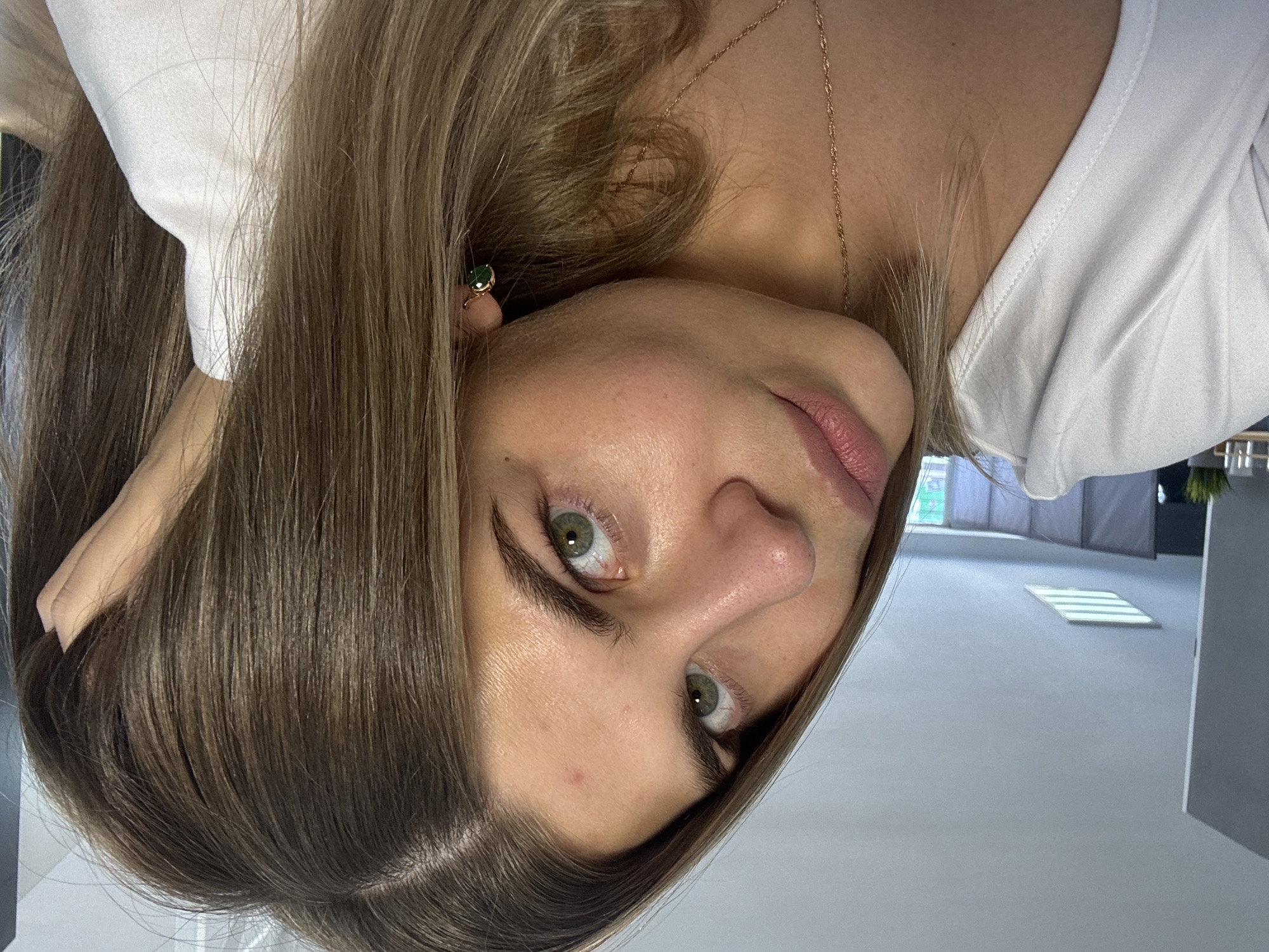 A close-up, upside-down, portrait-style image of a young woman with shoulder-length, highlighted brown hair wearing a white shirt and a gold necklace and earrings. She is indoors, likely in a room with a white floor and modern decor. The location is potentially Chaikovsky, Russia, based on the prompt's parameters. The woman has fair skin, green eyes, and slightly parted lips. She appears to be looking at the camera.