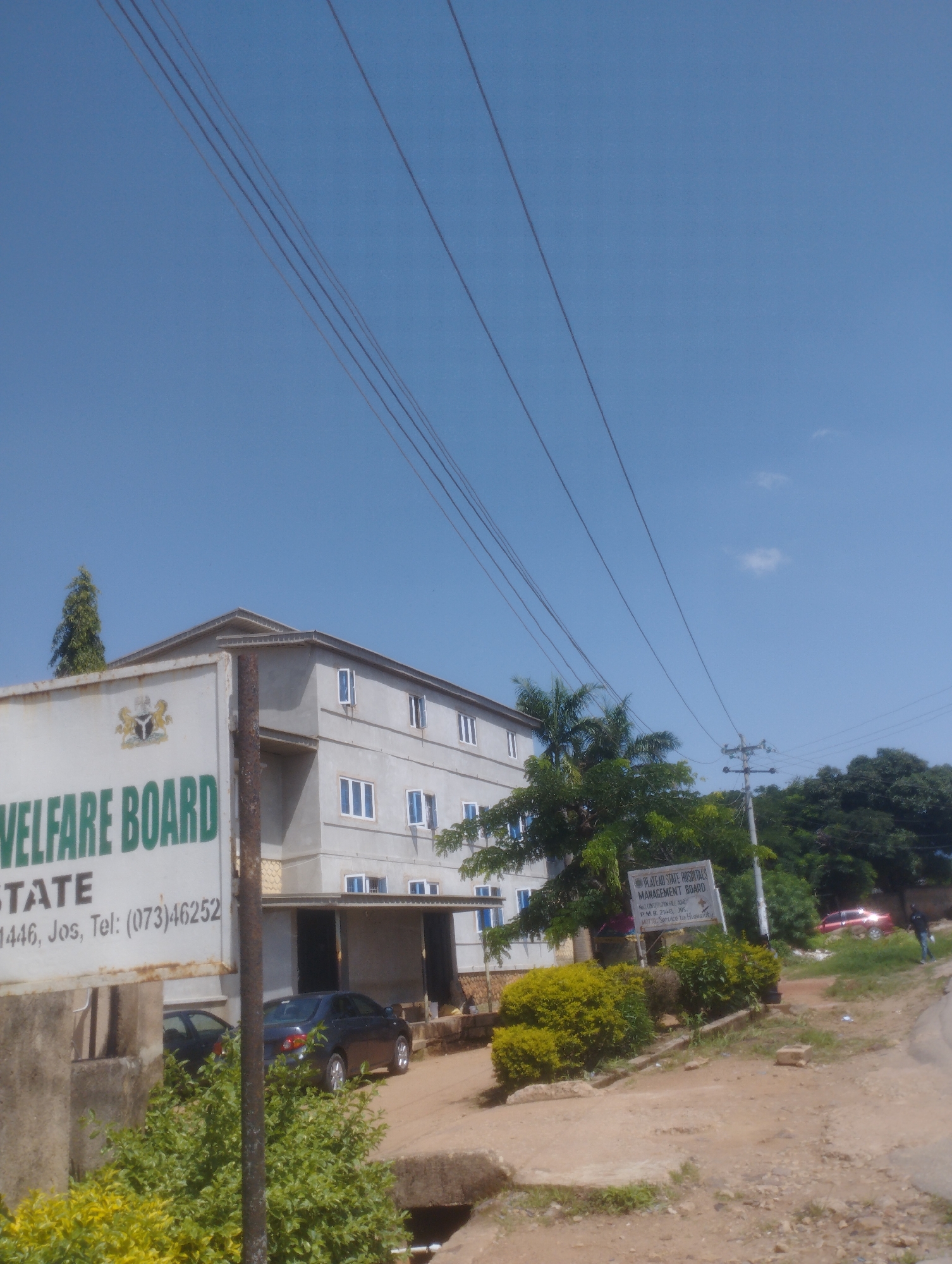 The image shows a low-angle view of a multi-story concrete building with a sign that reads "WELFARE BOARD STATE 1446, Jos, Tel: (073)46252". The building appears to be under construction or recently completed, with unpainted concrete walls and visible windows. Several power lines stretch across the clear blue sky, and a utility pole is visible to the right of the building. Green trees and shrubs are present in the foreground and around the building. A dark-colored sedan is parked in front of the building, and another car is partially visible behind it. Further down the road, a red car is parked and a person in a dark shirt and blue jeans is walking away from it. A second sign, partially obscured by trees, reads "PLATEAU STATE HOSPITALS MANAGEMENT BOARD". The scene is set in a relatively undeveloped area, likely on the outskirts of the city.