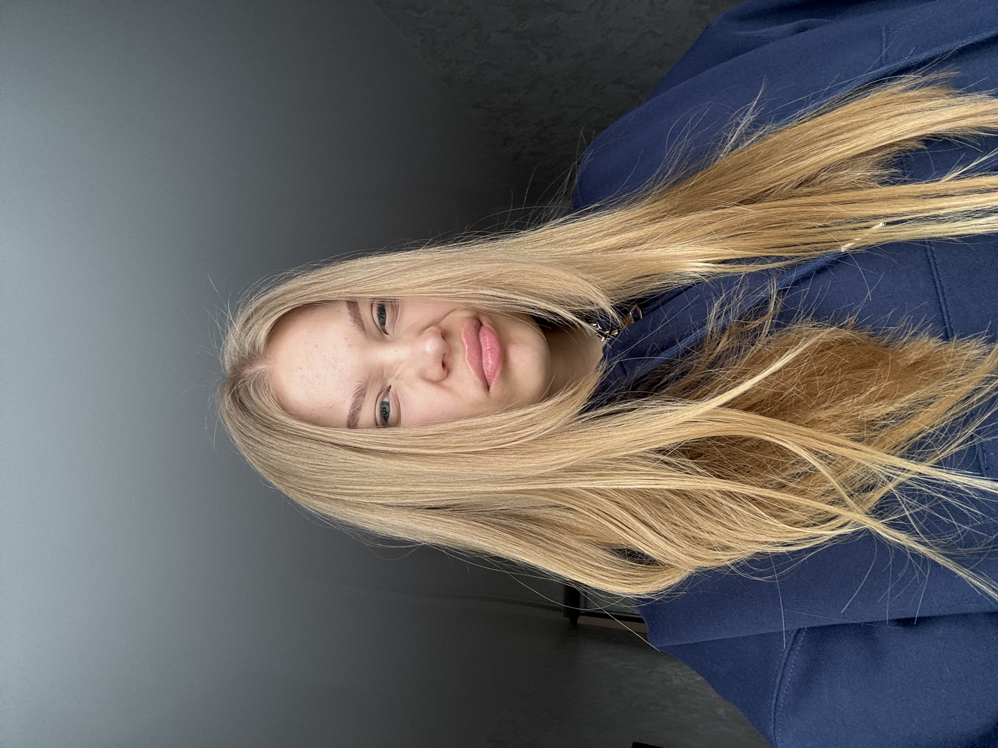A blonde-haired woman wearing a blue hoodie poses for a selfie. She is indoors, in front of a gray wall. No information confirms the location is in Chaikovsky, Russia.