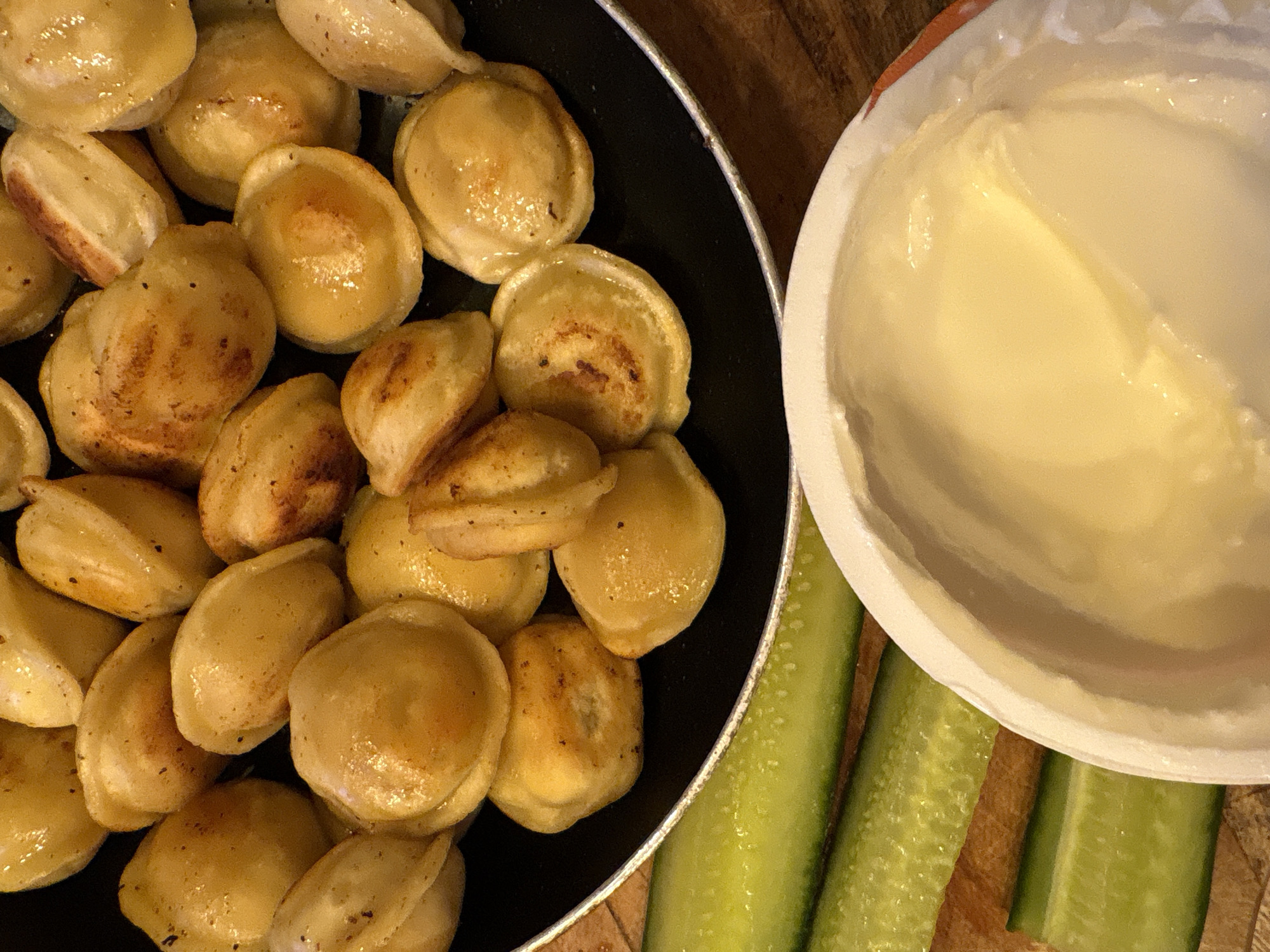 A close-up, overhead shot showcases a pan filled with golden-brown, pan-fried dumplings, likely pelmeni, on a dark surface. The dumplings appear slightly oily and some have browned, crispy edges. In the foreground, three cucumber sticks are arranged horizontally, and to the right, a white bowl contains a creamy, pale yellow substance, possibly sour cream or yogurt. The image is taken from a high angle, focusing on the food. No specific location in Krasnoyarsk, Russia, can be determined from the visual information provided.