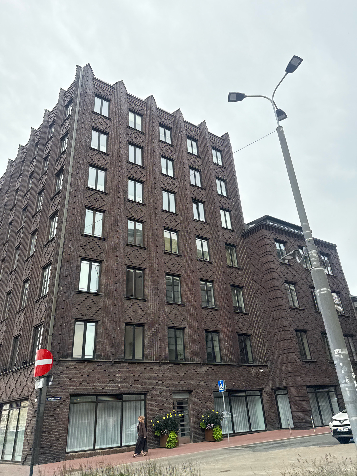The image shows a multi-story brick building with a distinctive Art Deco style, located in Tallinn, Estonia. The building features repeating diamond patterns in the brickwork on its facade and a stepped roofline. Several windows are visible, some with white curtains. In the foreground, a street lamp with three lights stands tall. A pedestrian walkway runs along the front of the building, and a woman in dark clothing is walking on it. A street sign indicating "Roosikrantsi" is visible. There is also a red and white prohibition sign on a pole. A white car is parked on the right side of the image. The sky is overcast.