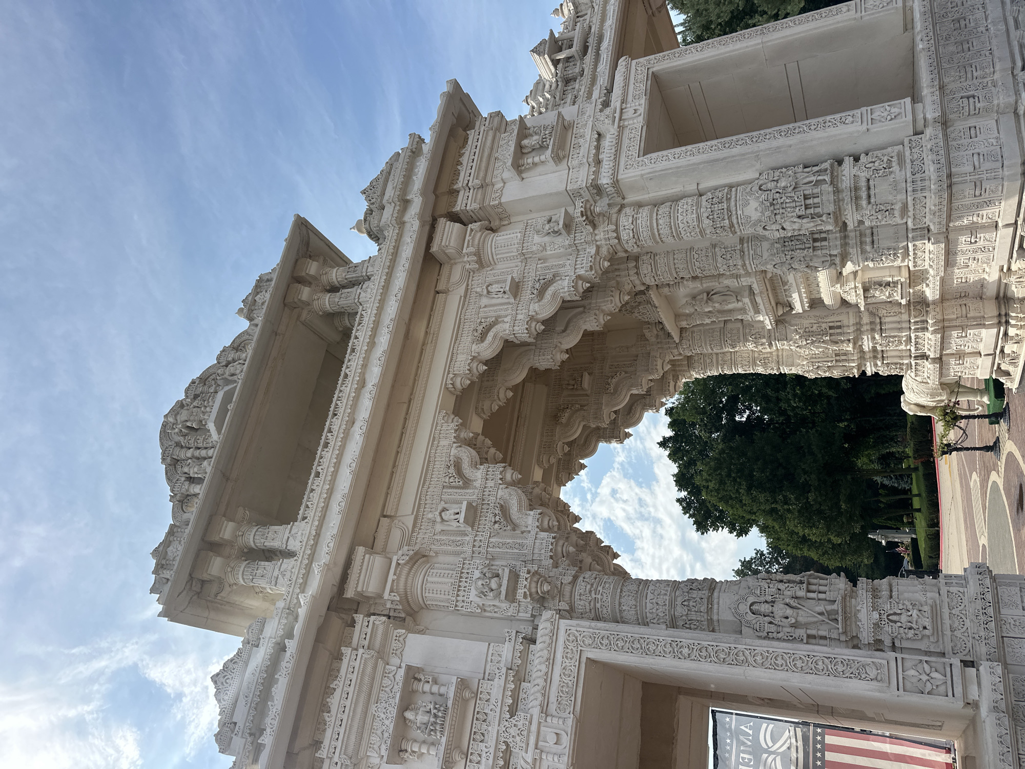 The image captures a close-up, low-angle view of a white, intricately carved stone archway, likely part of a temple structure. The archway is adorned with numerous small sculptures and decorative patterns. Above the arch, a tiered roof structure with similar carvings is visible against a bright blue sky with scattered wispy clouds. Green trees and foliage are visible through the archway and to the right of the structure. On the left side of the archway, a banner with an American flag motif is partially visible. In the foreground, to the right, a portion of a white elephant sculpture is visible, adorned with decorative coverings. The scene appears to be outdoors, possibly in a garden or park setting, given the presence of trees and manicured grounds. The specific location is identified as the Shri Swaminarayan Mandir in Bartlett, Illinois, United States of America.