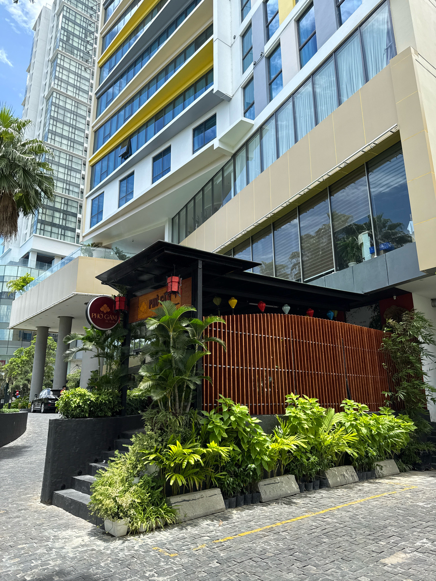 A low-angle shot captures the exterior of a modern building and a restaurant in Nha Trang, Vietnam. The building is tall and features a facade of white, yellow, and grey elements with numerous windows. In front of the building, a restaurant with a black roof and bamboo-like wooden siding is visible. A circular sign with Vietnamese text and the words "PHO GAMA" is attached to the restaurant's entrance. The restaurant is adorned with colorful paper lanterns hanging from the roofline. Lush green plants and potted vegetation surround the entrance and line the walkway. The ground is paved with dark grey cobblestones, and a yellow line is painted on the surface. A dark-colored car is parked on the left side of the frame, partially obscured by a black retaining wall and a set of stairs. The sky is visible above, with some clouds present.