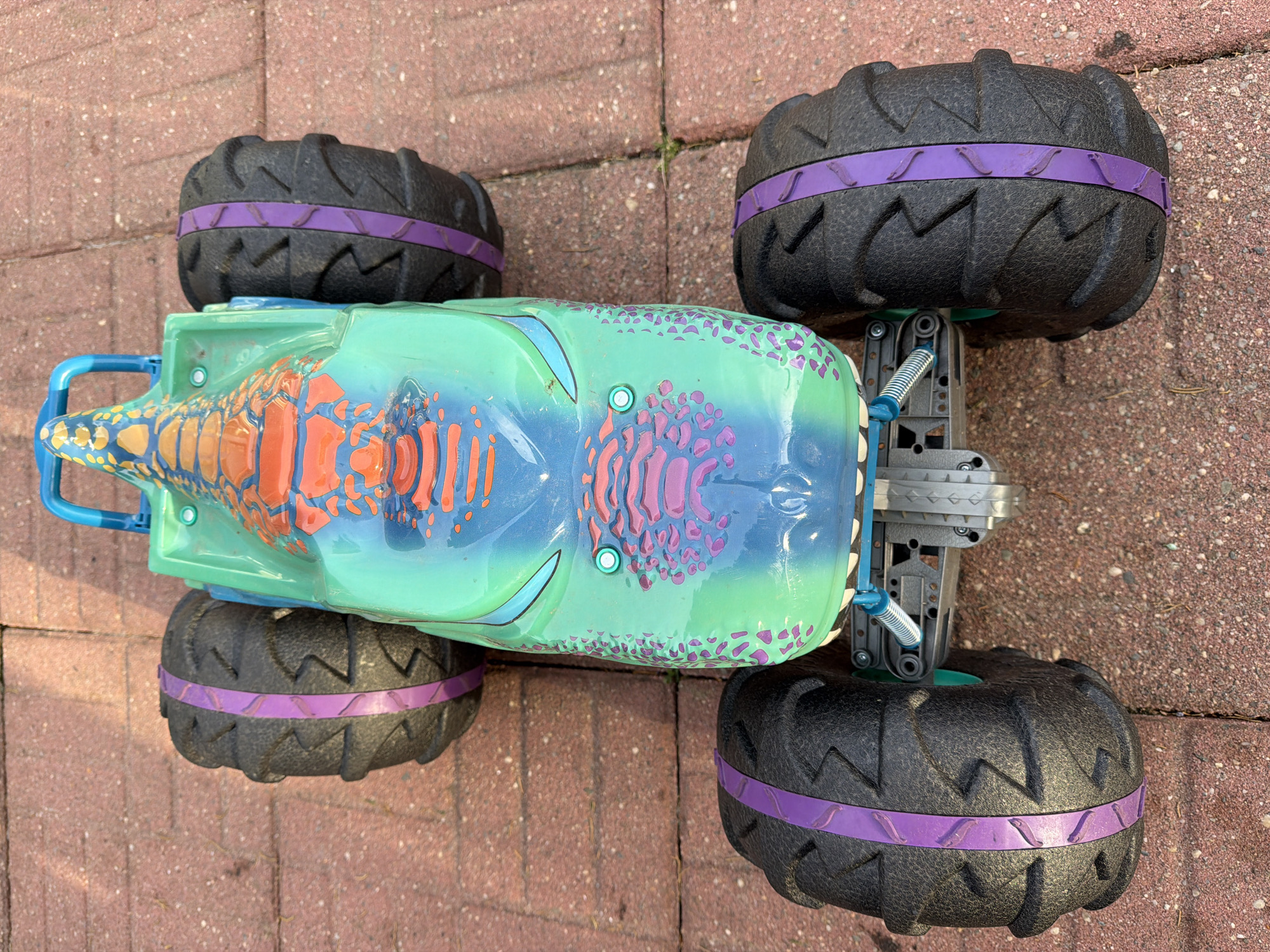 A high-angle shot shows a toy monster truck with large black tires with purple accents on a brick-paved surface. The monster truck has a predominantly teal and blue body with orange and pink tribal markings. The truck appears to be stationary. The environment suggests an outdoor setting in Riverhead, United States of America.