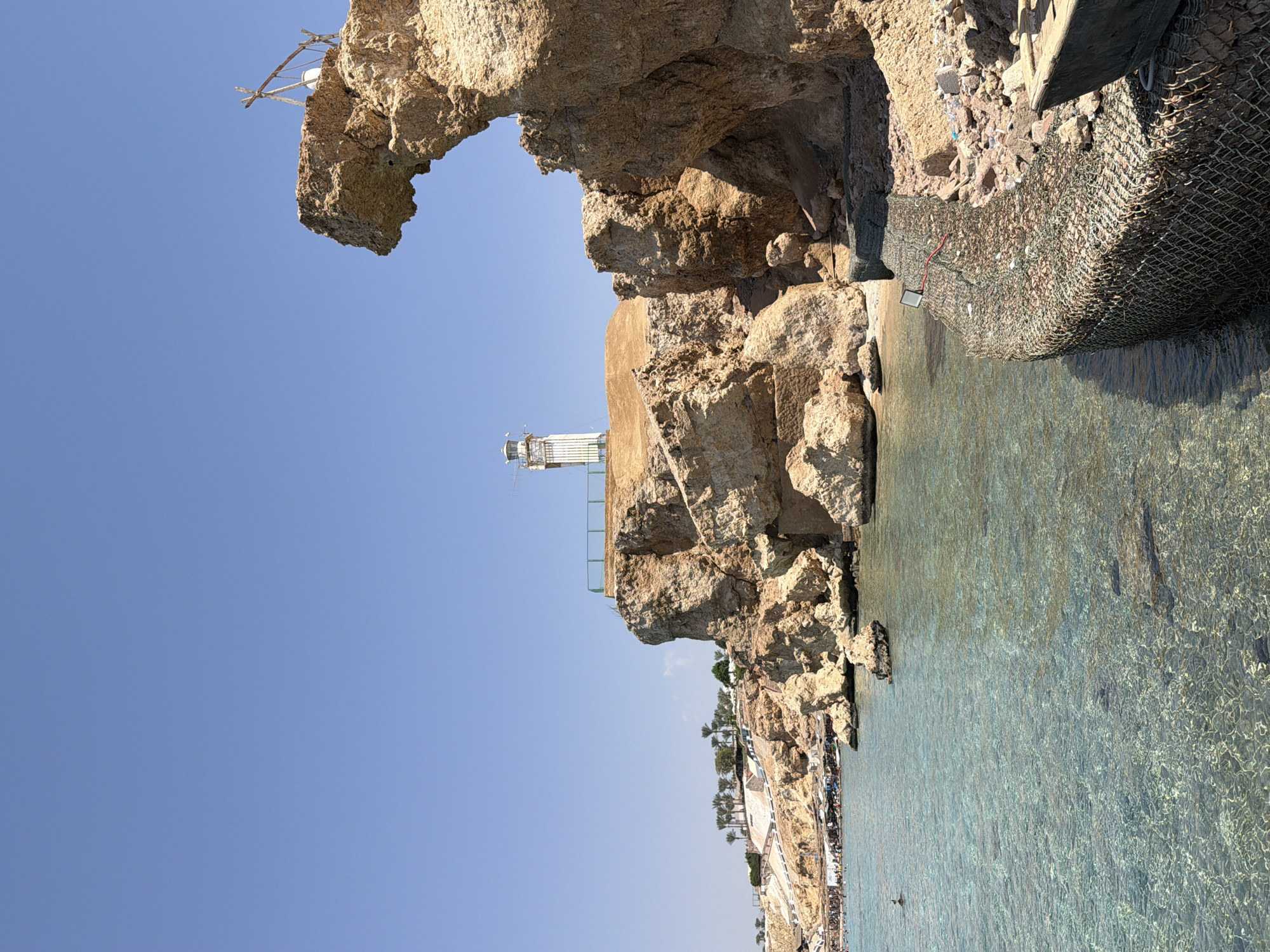 The image depicts a coastal scene in Sharm El Sheikh, Egypt, featuring a rocky coastline with a lighthouse perched on a cliff. The sea in the foreground is clear and shallow, revealing rocks beneath the surface. To the left, a beach is visible with numerous people sunbathing and swimming. Palm trees and structures line the cliff face in the background, suggesting a resort or populated area. A wire mesh structure, possibly a barrier or erosion control, is prominent in the lower right of the frame, with a small boat resting on it. The sky is clear and blue, indicating a sunny day.