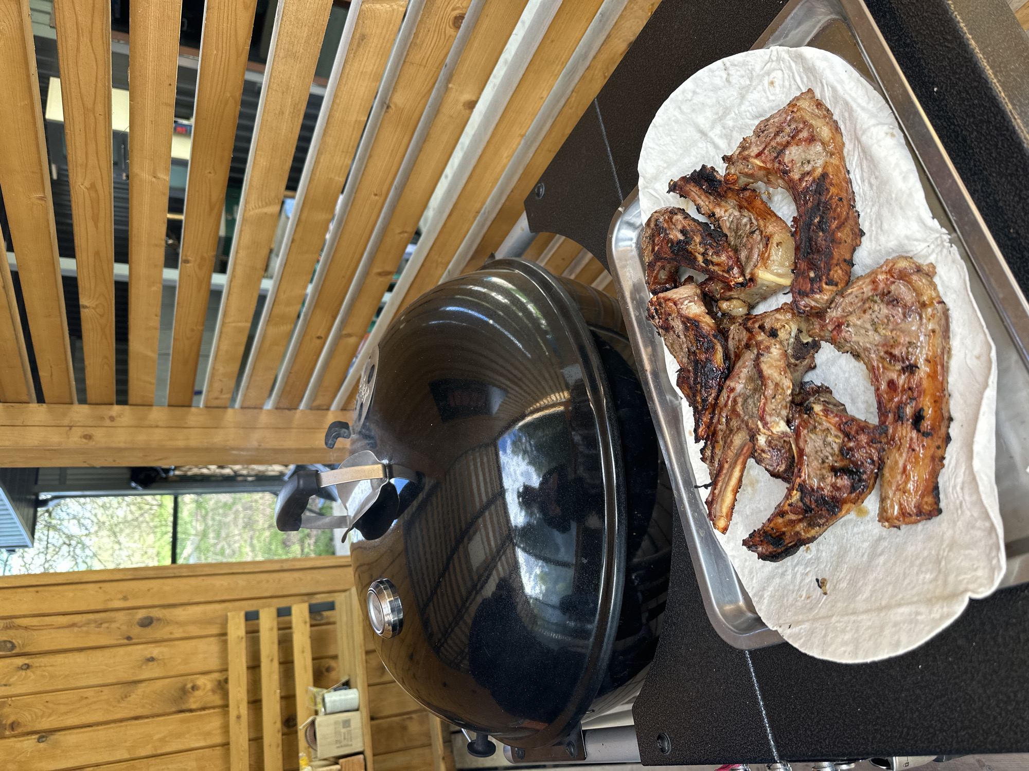 A close-up, angled shot captures a black grill situated on a wooden deck with a tray of cooked lamb chops placed on a paper towel, within what appears to be an outdoor covered grilling area in Прикамский, Russia. The wooden structure features both solid horizontal panels and open-slatted sections above. The grill, reflecting the surrounding environment on its glossy surface, is the central element, while the lamb chops, displaying grill marks, are arranged on a metal tray. The background reveals glimpses of greenery visible through the slatted portions of the wooden structure.