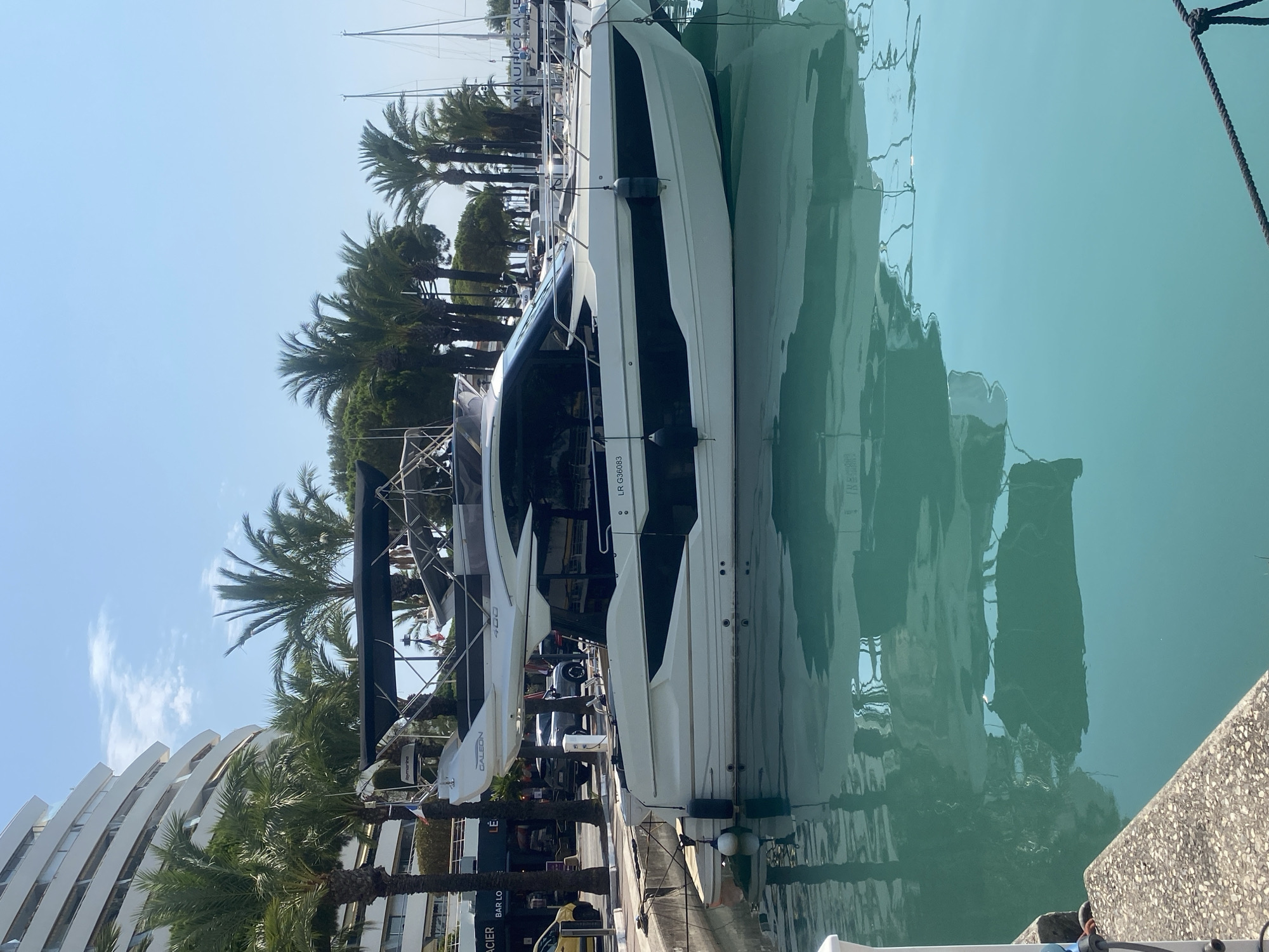 In Villeneuve-Loubet, France, a white motor yacht named "GALEON" with a black trim is docked in a marina. The yacht is positioned parallel to a concrete walkway, with its reflection visible in the clear turquoise water. Palm trees and a modern, white multi-story building line the background, suggesting a resort or residential area. The sky is clear and blue, indicating a sunny day. Several other boats and masts are visible in the distance, along with a portion of a street with a yellow vehicle. The yacht has a sleek design, with a dark-tinted windshield and a hardtop. Fender lines are attached to the yacht, securing it to the dock.