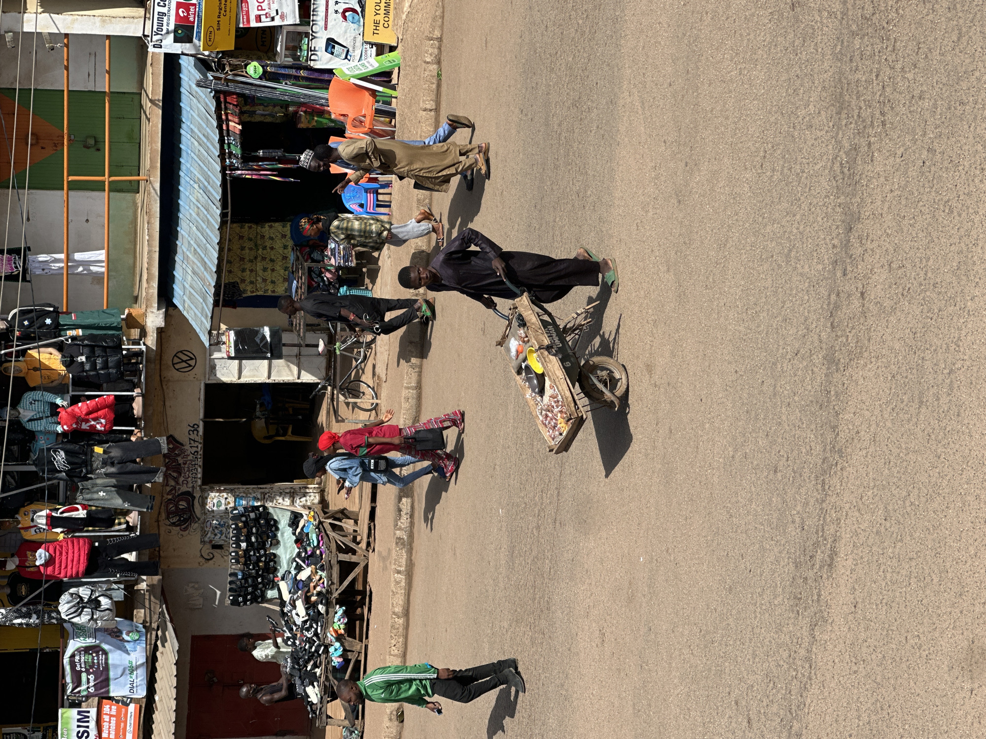 The image depicts a street scene in Bukuru, Nigeria, likely a market area. In the foreground, a young man in a dark robe pushes a wheelbarrow filled with various items, including what appear to be food products. To the left, a man in a green jacket walks away from the camera, holding a phone. Further back, two women, one in a blue denim jacket and the other in a red headscarf and skirt, are walking and looking at their phones.
Behind them, a stall displays numerous pairs of sandals. In the mid-ground, several men and women are present near market stalls. Above, a clothing shop displays mannequins wearing jackets and other garments. The environment is urban, with buildings and a paved street. The lighting suggests it is daytime.