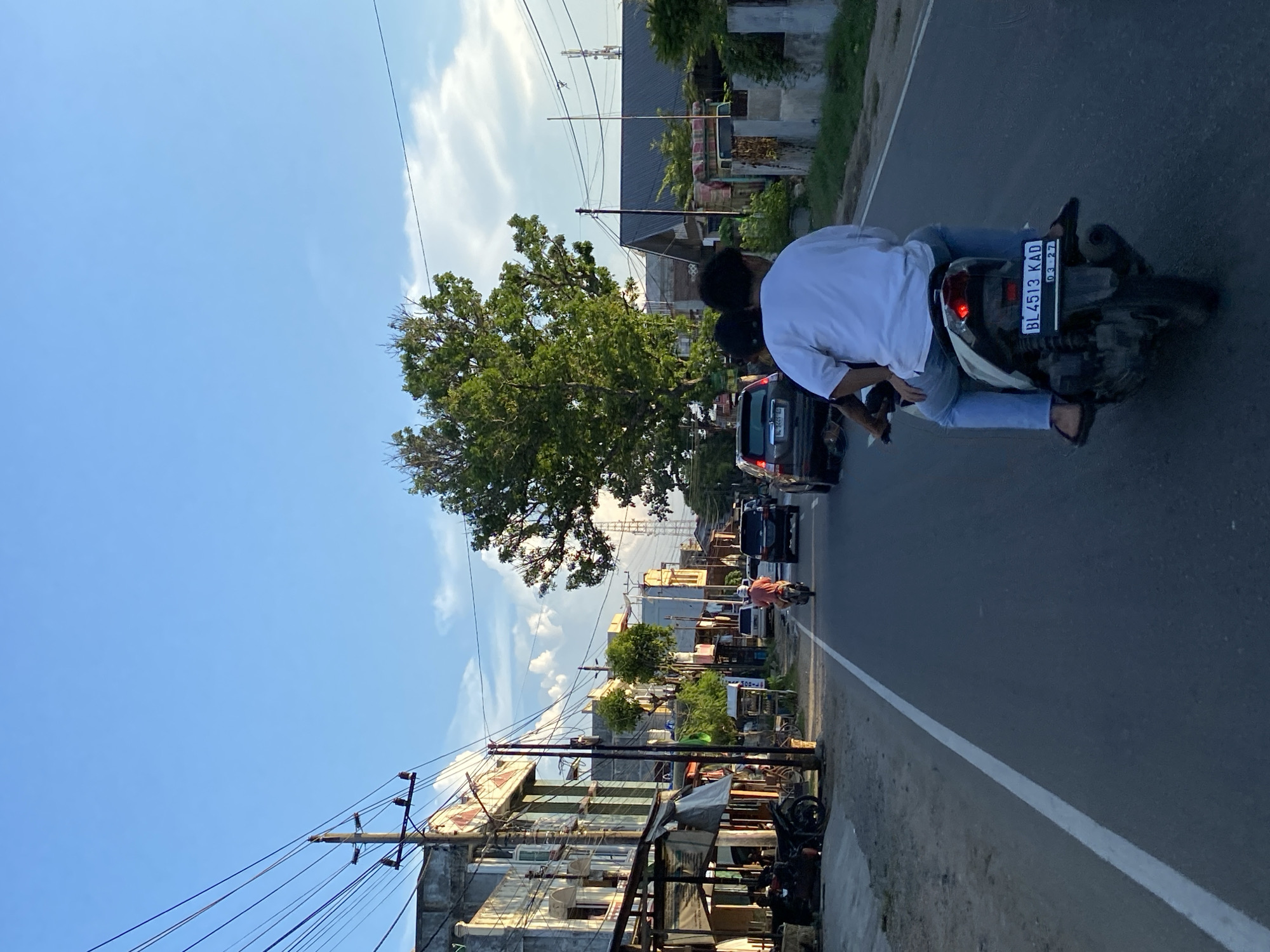 The image shows a street scene in Lhokseumawe, Indonesia, likely during late afternoon or early evening, given the lighting. The perspective is from behind a motorcycle carrying two people, a driver and a passenger, both appearing to be young adults. The driver is wearing a white t-shirt and blue jeans, and the passenger is wearing a white t-shirt and blue jeans. They are riding on a paved road with a white dividing line.
The street is lined with buildings on the left, some appearing to be shops or small businesses with awnings and signs. Several motorcycles are parked on the side of the road. On the right side of the road, there are more buildings, fences, and dense foliage, including a large tree prominently in the middle ground.
Several vehicles are visible on the road: a black SUV in front of the motorcycle, another black SUV further ahead, and a white car. A person on a separate motorcycle, wearing an orange shirt, is also visible in the distance. Utility poles with numerous wires crisscross the sky above the street. The sky is a clear blue with some scattered white clouds. The overall impression is a typical urban or suburban street scene in Indonesia.