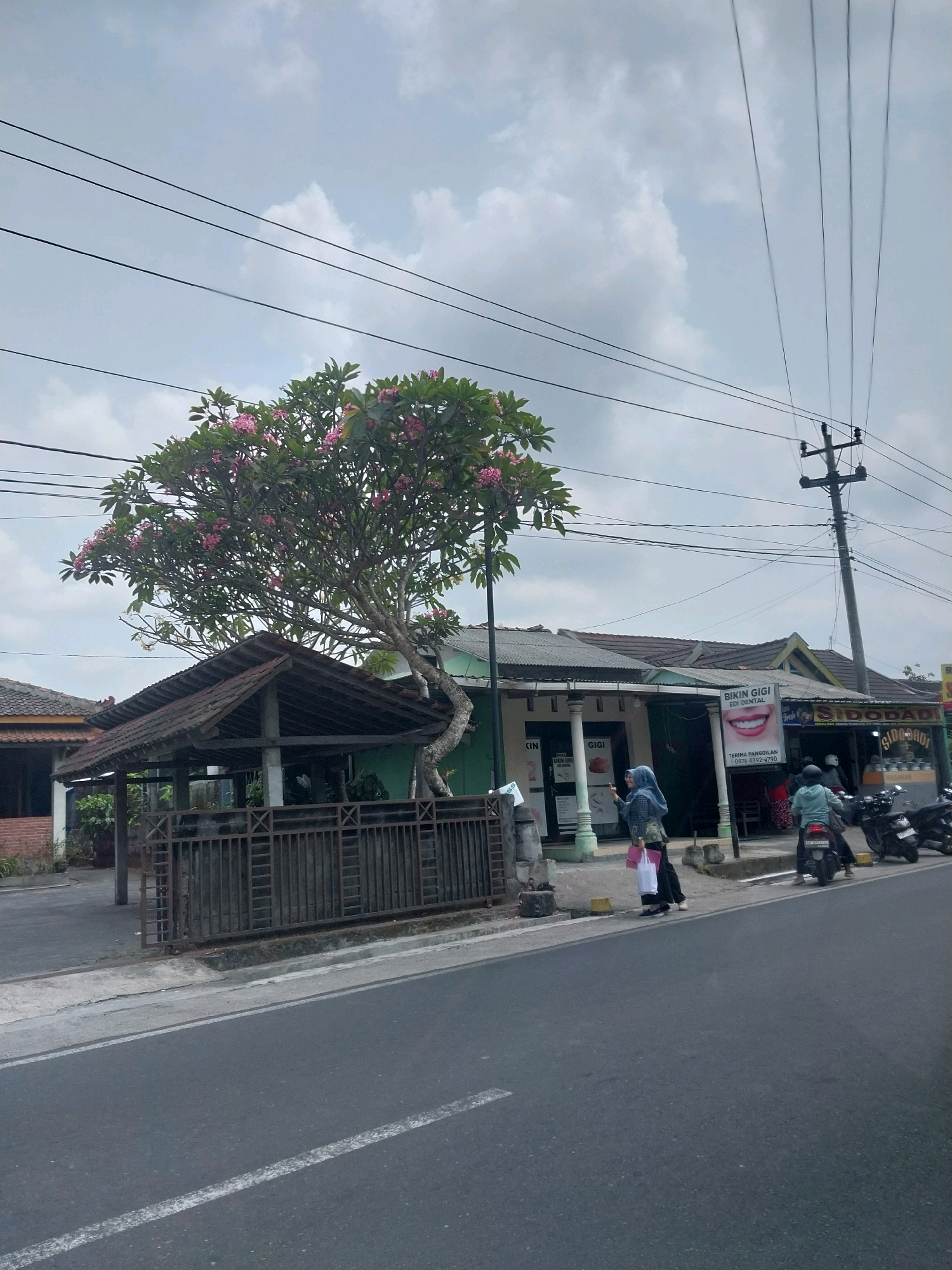The image captures a street scene in Tirtomartani, Indonesia. In the foreground, a gray asphalt road with a white dividing line runs diagonally across the frame. To the left of the road is a concrete sidewalk and a metal fence. Behind the fence, a small, open-air structure with a tiled roof is visible, along with a tree bearing pink flowers. Further back, a row of buildings lines the street. One building prominently displays a sign for "BIKIN GIGI" (Make Teeth) and "EDI DENTAL," indicating a dental clinic. Another building to its right has a sign that reads "SIDODADI." Several people are present: two women are walking on the sidewalk near the dental clinic, one carrying a pink bag, and another in a blue headscarf. A person is seated at a table outside the SIDODADI establishment. In the middle ground, two motorcycles are parked, and one is being ridden away from the camera. Overhead, multiple power lines are suspended from utility poles, crisscrossing the cloudy sky. The overall scene suggests a typical commercial street in a populated area.