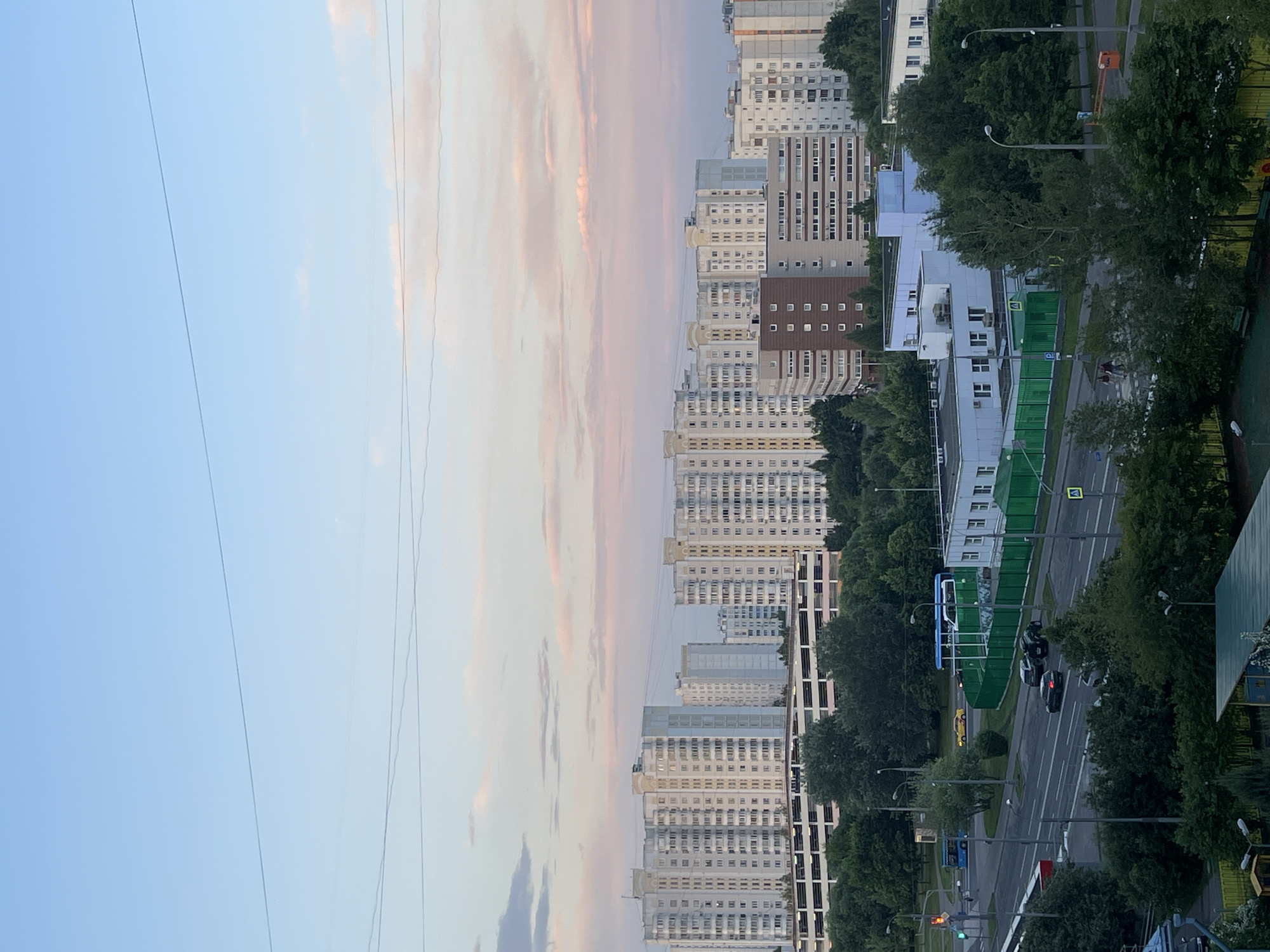The image depicts a residential urban landscape at dusk, likely in a Moscow, Russia suburb, given the architectural style of the buildings. In the background, a series of mid-rise apartment buildings, typical of Soviet-era mass housing, dominate the skyline. These buildings are predominantly light-colored, with numerous windows and balconies. A multi-level parking garage is situated in front of some of these buildings.
In the mid-ground, a road runs through the scene. Several vehicles are visible, including a blue bus, a yellow car, and at least two dark-colored sedans. A pedestrian crossing with a traffic light is present, indicating an intersection. Adjacent to the road, a long green fence encloses an area, possibly a school or administrative complex, with low-rise white buildings behind it. Trees and greenery are interspersed throughout the scene, softening the urban environment.
The sky above displays a gradient of colors, transitioning from a light blue at the top to soft pink and orange hues near the horizon, characteristic of a sunset. Several thin power lines are stretched across the sky, a common urban feature. The overall impression is one of a functional, albeit somewhat monotonous, residential area in the late evening.