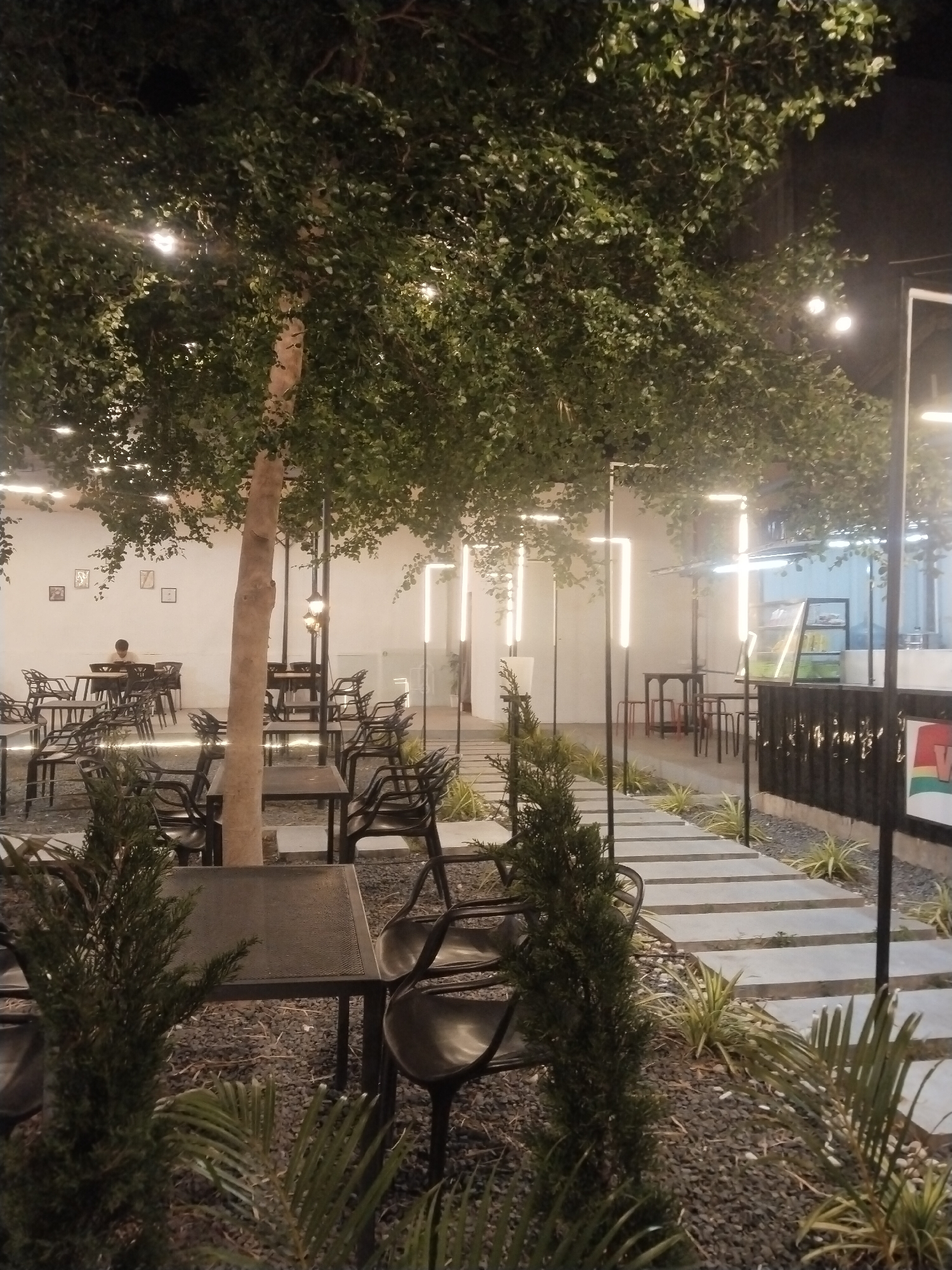 The image depicts an outdoor dining area, likely a restaurant or cafe, at night. The location is possibly Lhokseumawe, Indonesia. The scene is lit by string lights strung through trees and vertical, rectangular light fixtures. Tables and chairs, mostly black, are scattered throughout the area, some arranged under trees. There is one visible patron seated at a table in the background. A pathway made of rectangular concrete slabs cuts through the area. Gravel, plants, and small trees are around the tables and along the pathway. The overall ambience suggests a relaxed, al fresco dining setting.
